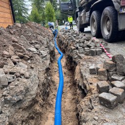 eFLOW - Niebieska rura karbowana w wykopie, prawdopodobnie pod kable, z pracownikami i ciężarówką w tle. Widoczne kostki brukowe i ziemia.