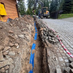 eFLOW - Wykop z niebieskimi rurami w ziemi, przygotowany pod ułożenie kabli. Na górze koparka i kostka brukowa. Żółta skrzynka gazowa na drewnianym budynku w tle.