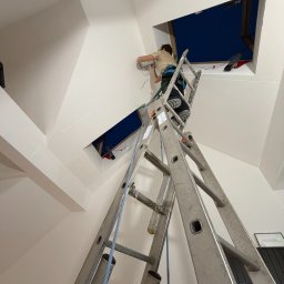 eFLOW - Elektryk na drabinie montuje instalację w otworze w skośnym suficie. Widoczne przewody i laserowy wskaźnik poziomu. Nietypowy kształt otworu okiennego.