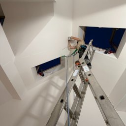 eFLOW - Elektryk na drabinie montuje osprzęt w skosie pod oknem dachowym, zabezpieczony uprzężą. Widoczne zielone linie lasera wyznaczające poziom. Białe ściany i sufit.