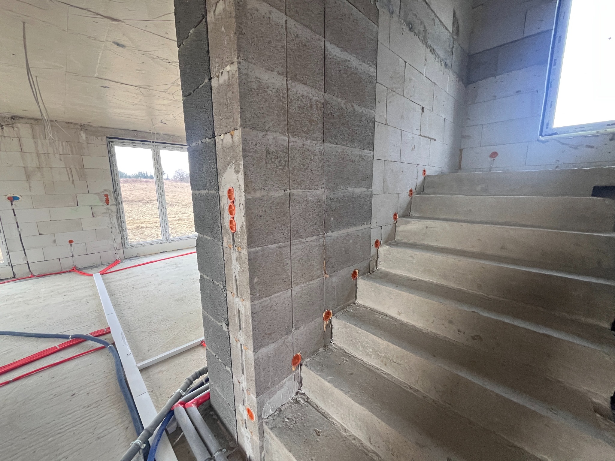 Surowy stan budynku w trakcie prac: widoczne kable elektryczne w ścianach z betonu komórkowego i rury na podłodze. Schody betonowe w tle.