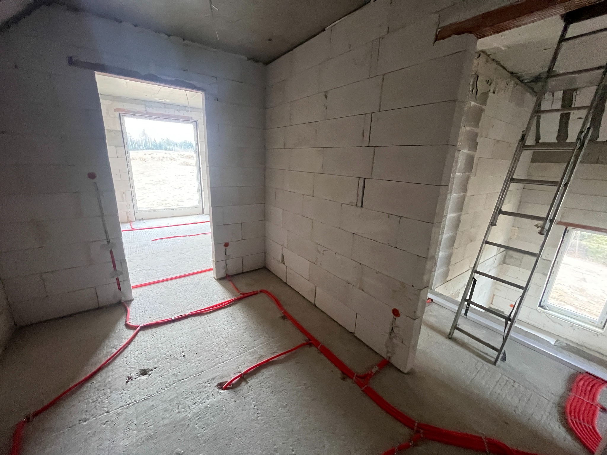 Czerwone rury ogrzewania podłogowego ułożone w surowym pomieszczeniu z białych bloczków. Widoczna drabina i okno z widokiem na zewnątrz. Etap prac instalacyjnych.