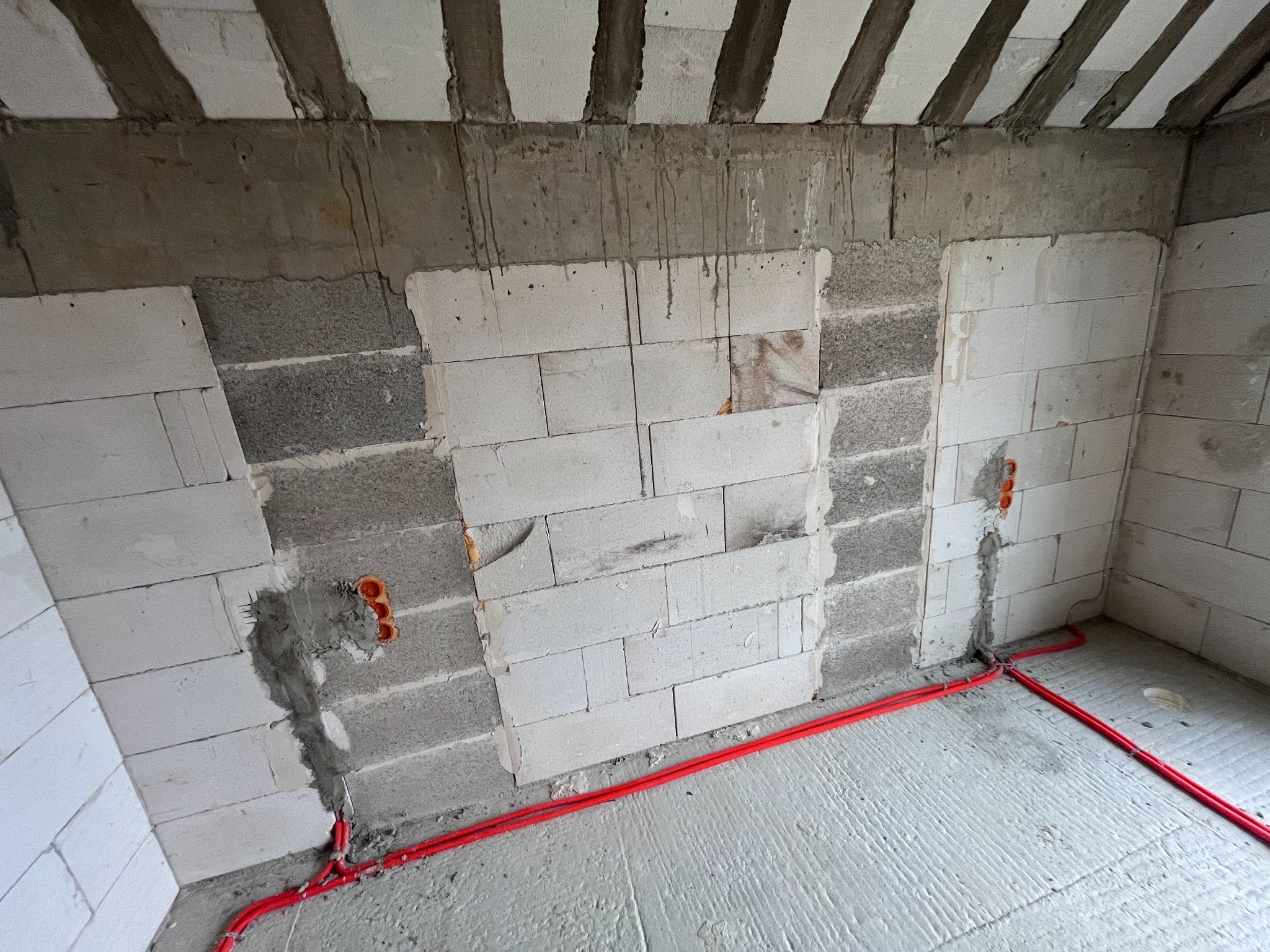 Czerwone kable elektryczne ułożone na posadzce w surowym wnętrzu z białymi ścianami z betonu komórkowego i widocznymi puszkami instalacyjnymi.