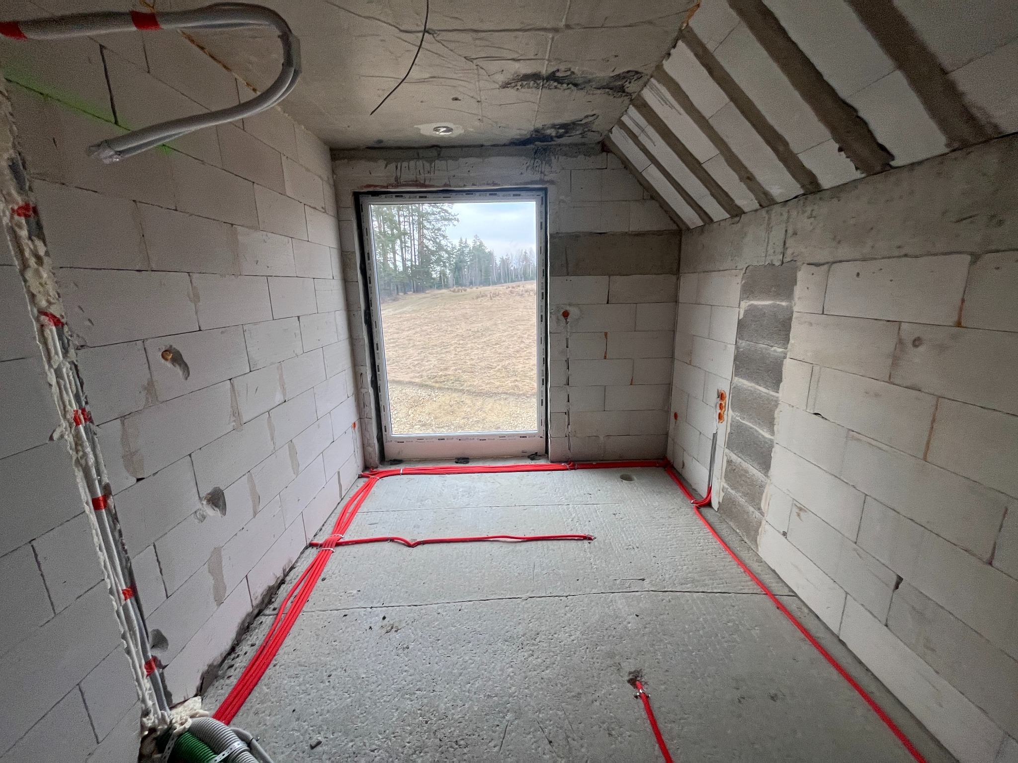 Surowy pokój z widokiem na zewnątrz przez okno, z widocznymi czerwonymi kablami elektrycznymi ułożonymi wzdłuż ścian i podłogi, oraz szarymi rurami przy suficie.