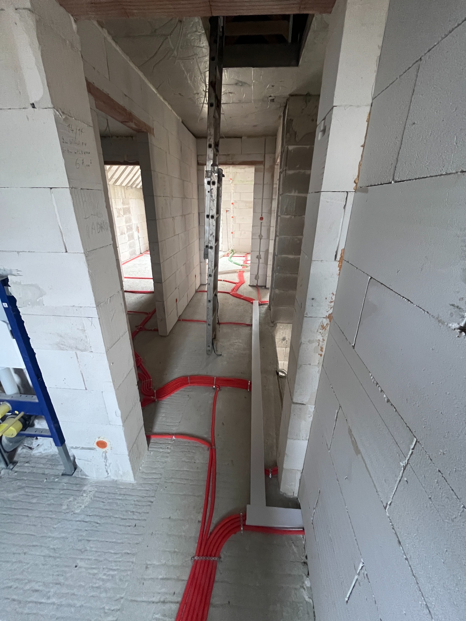 Wnętrze w trakcie budowy z widocznymi czerwonymi rurami instalacji ogrzewania podłogowego, białymi ścianami z betonu komórkowego i drabiną opartą o strop.