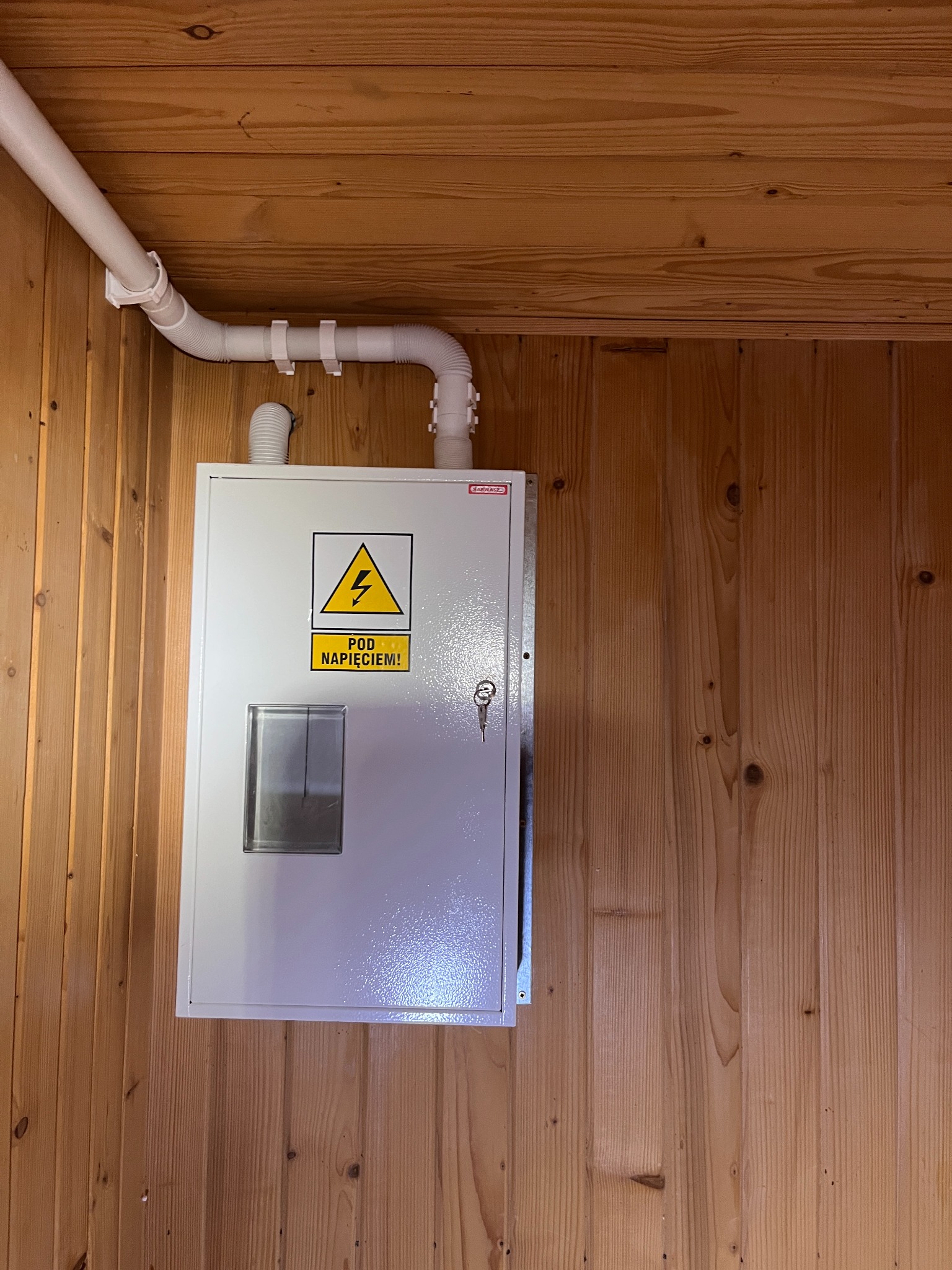Biała skrzynka elektryczna z ostrzeżeniem 'Pod Napięciem!' na drewnianej ścianie, z widoczną rurą ochronną. Widok z dołu, oświetlenie naturalne.
