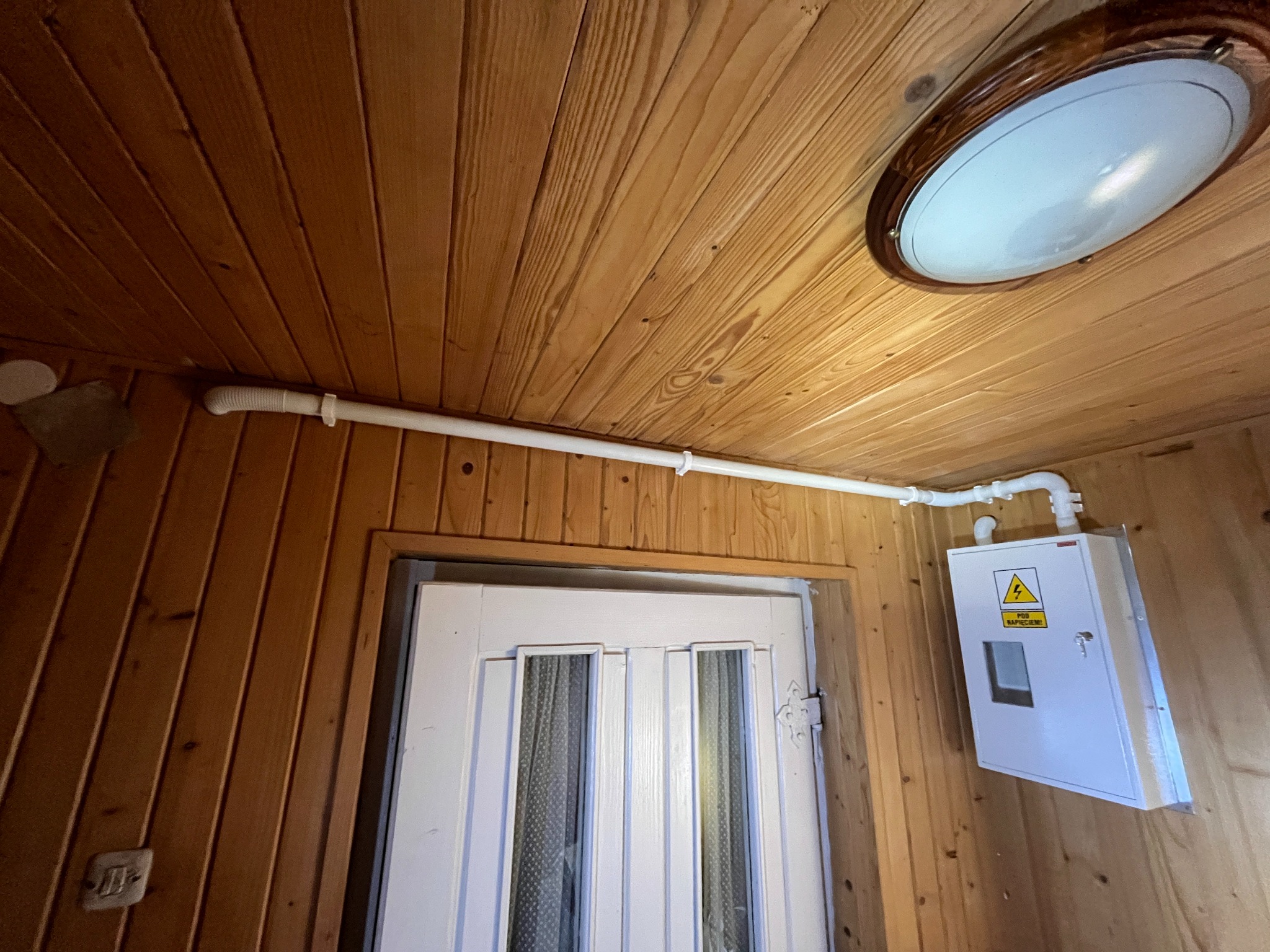Biała skrzynka elektryczna z żółtym ostrzeżeniem na drewnianej ścianie, widoczna biała rura instalacyjna i lampa sufitowa w drewnianym wnętrzu.