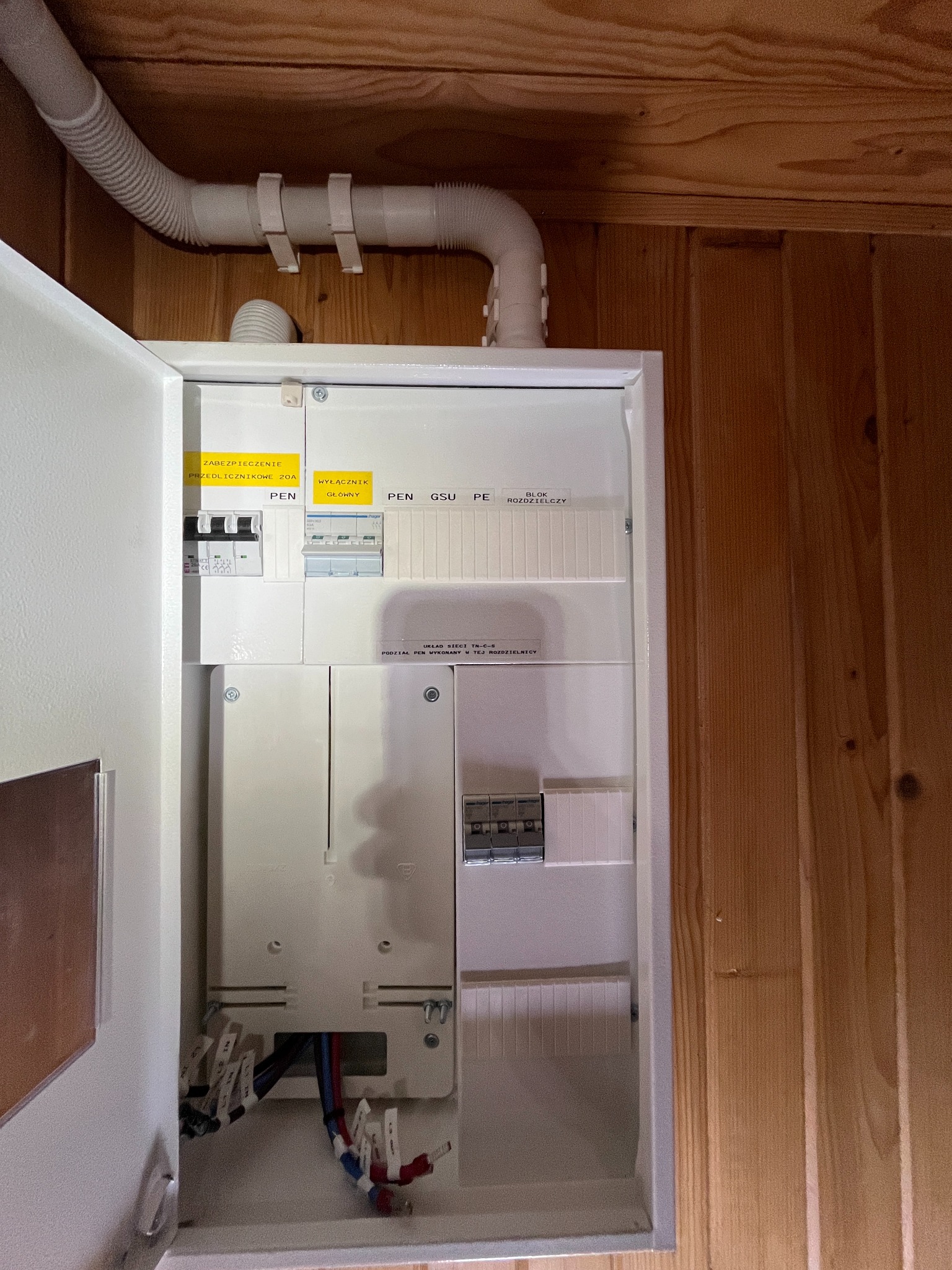 Otwarta, biała skrzynka elektryczna z widocznymi przewodami i bezpiecznikami, zamontowana na drewnianej ścianie. Rury ochronne nad skrzynką.
