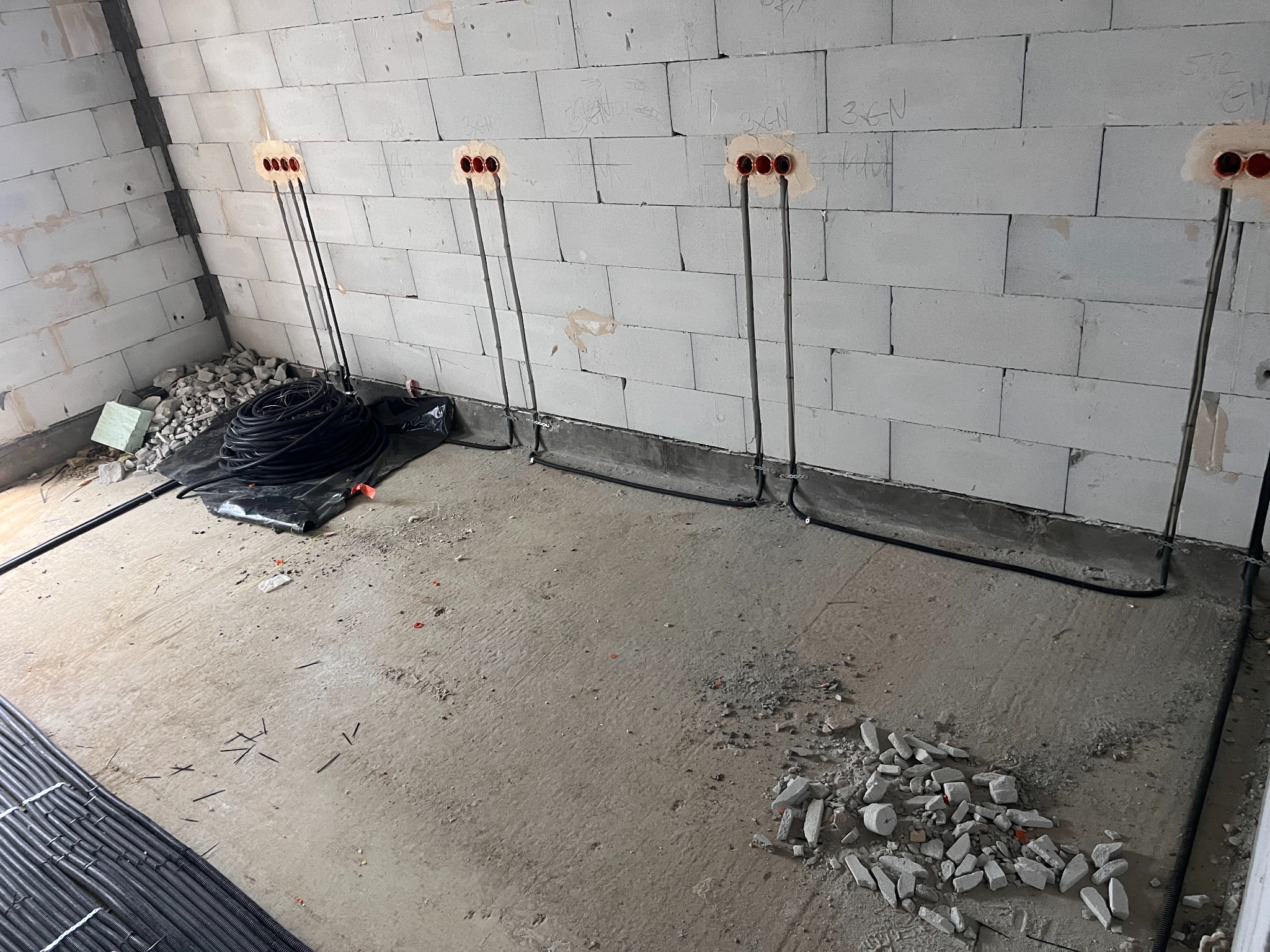 Surowy stan: instalacja elektryczna w trakcie montażu w nowym budynku. Widoczne przewody w ścianach z pustaków i zwój kabli na podłodze.