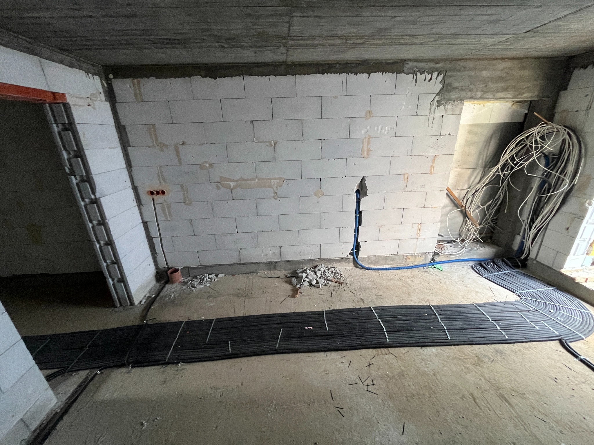 Stan surowy: przewody elektryczne ułożone na podłodze, instalacja w ścianie z białego pustaka, widoczne kable i rury. Przygotowanie do wylewki.