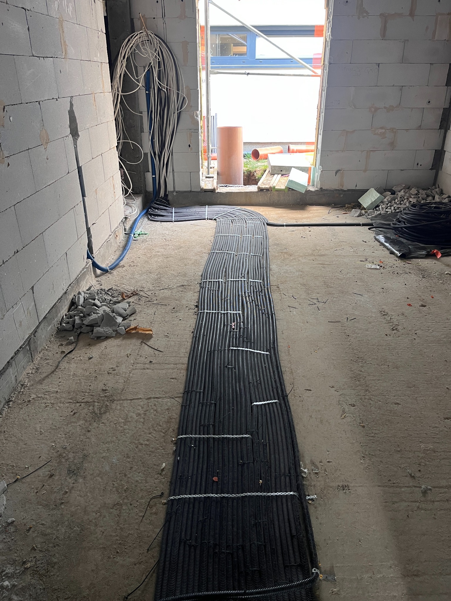 Kable elektryczne ułożone równolegle na podłodze w surowym wnętrzu budynku w trakcie budowy. Widoczne białe kable zwisające ze ściany i gruz.