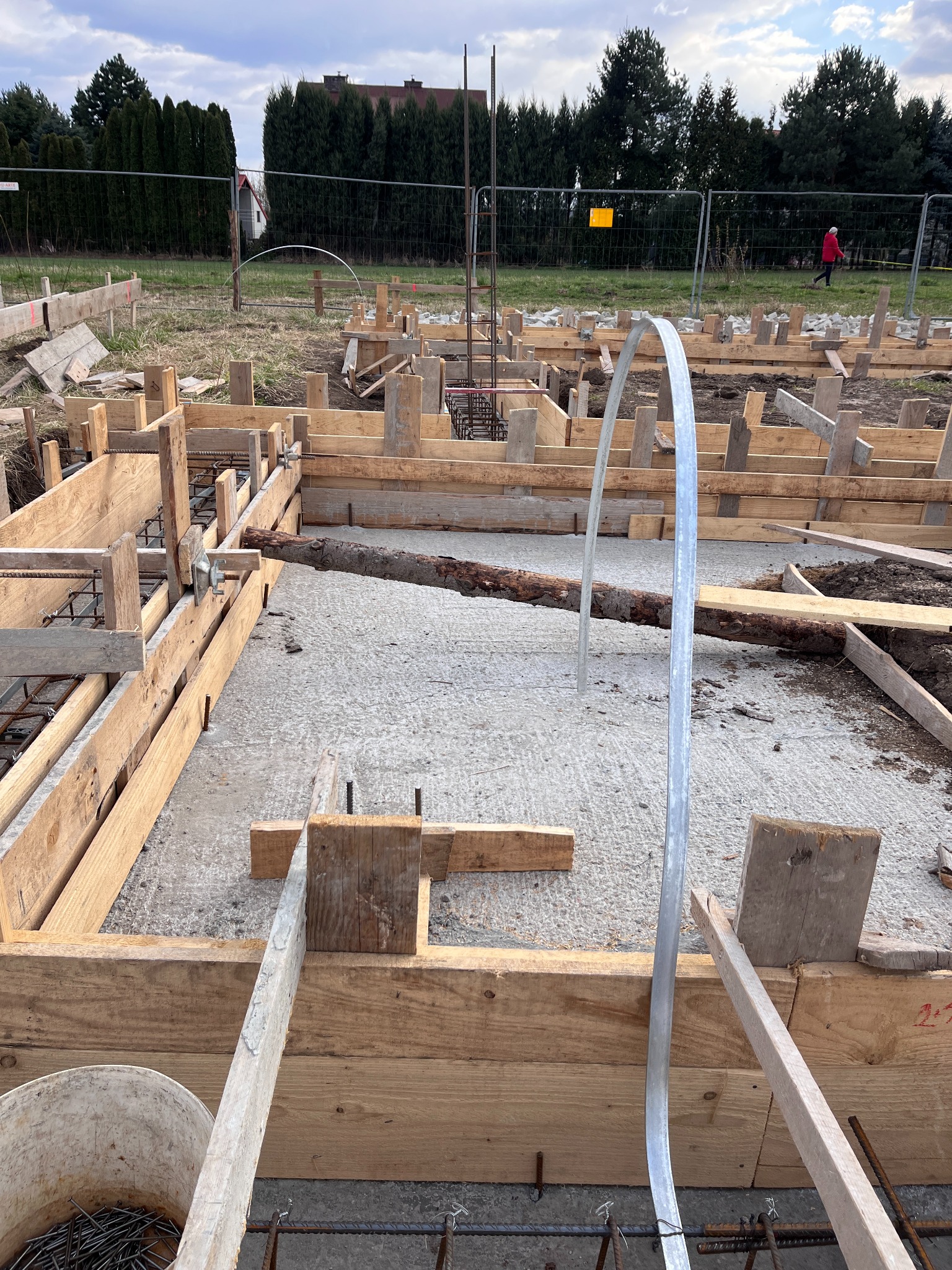 Szczegółowe ujęcie fundamentów domu w trakcie budowy: drewniane szalunki, zbrojenie stalowe, częściowo wylany beton. W tle widoczne drzewa i ogrodzenie.