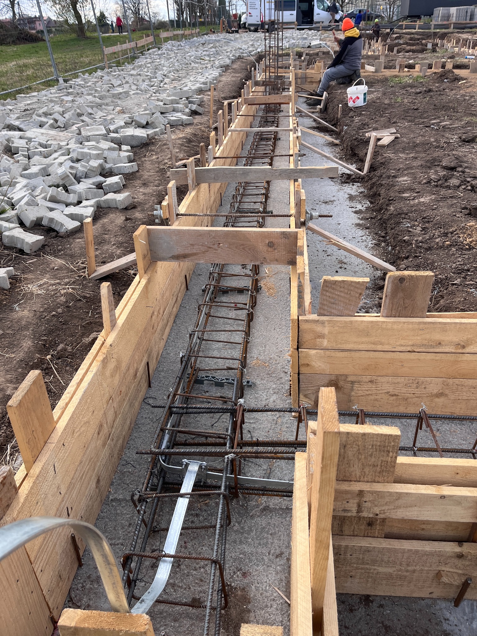 Wykop zbrojony stalą i szalunkiem z desek, przygotowany do wylania fundamentów. W tle pracownik w pomarańczowej czapce i sterta kostki brukowej.