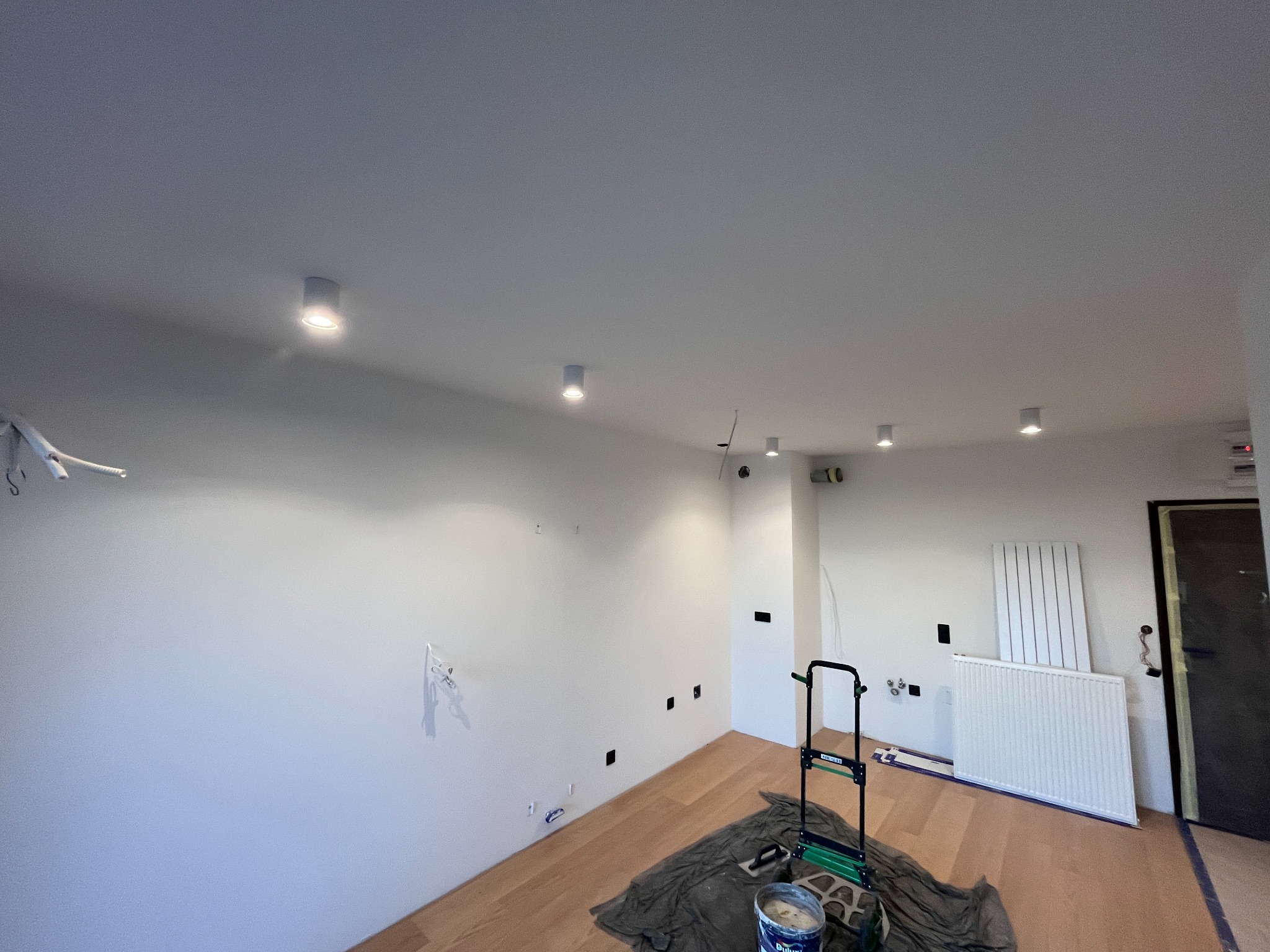 Wnętrze po malowaniu ścian na biało, widoczne nowe lampy sufitowe, zakryta podłoga i drabina malarska. Świeżo wykończone gniazdka elektryczne.