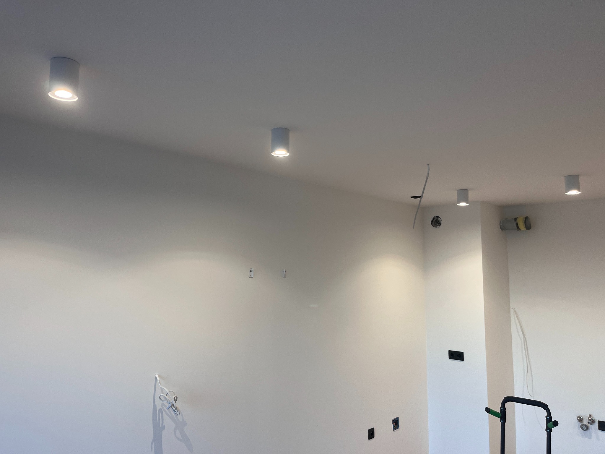 Nowoczesne, białe lampy sufitowe typu downlight, równomiernie rozmieszczone w jasnym pomieszczeniu. Widoczne kable i puszki instalacyjne, sugerujące etap wykończeniowy.