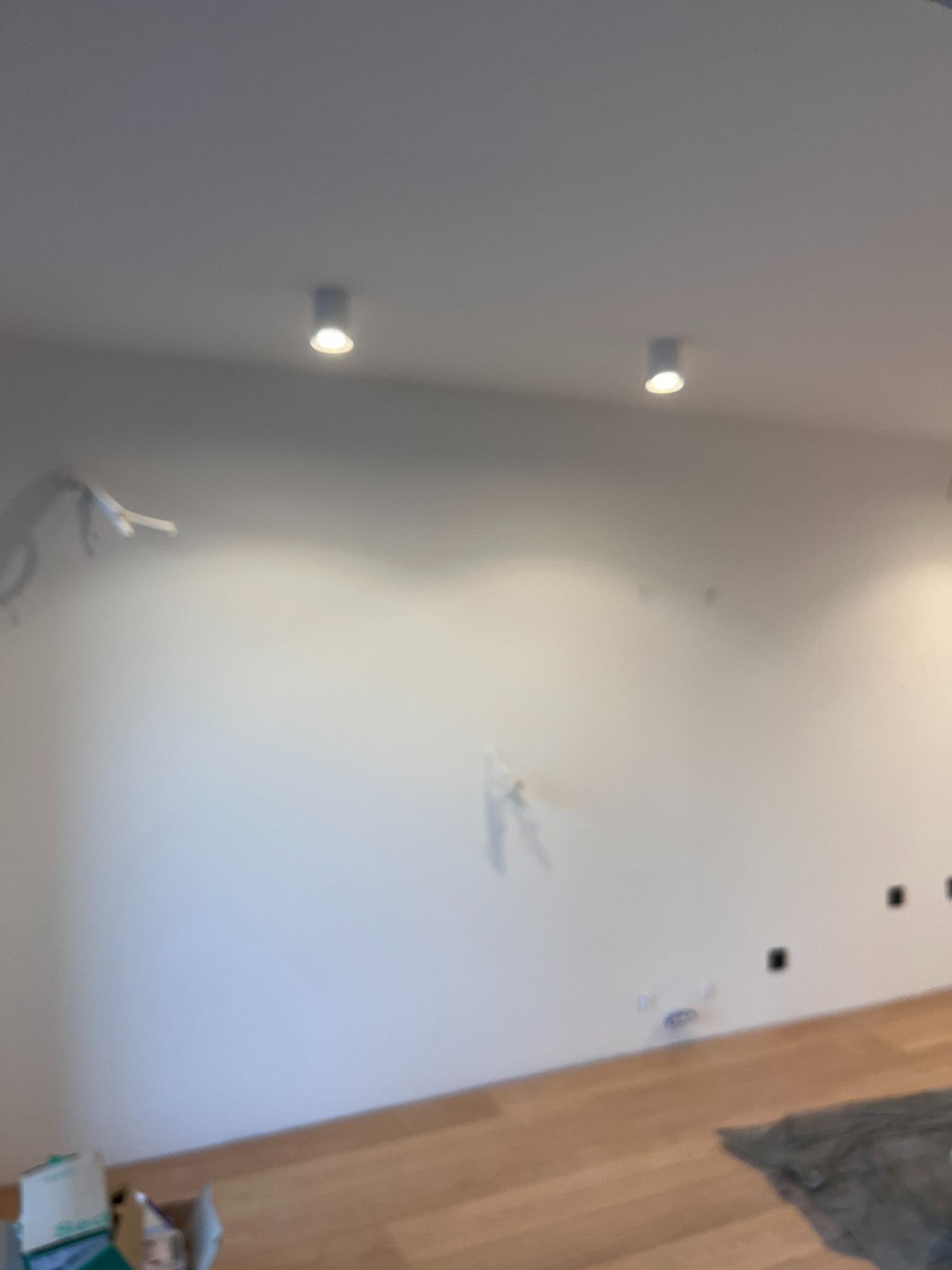 Biała ściana z dwoma punktowymi lampami sufitowymi. Widoczne wystające kable, puszki elektryczne i fragment podłogi z parkietu. Etap prac wykończeniowych.