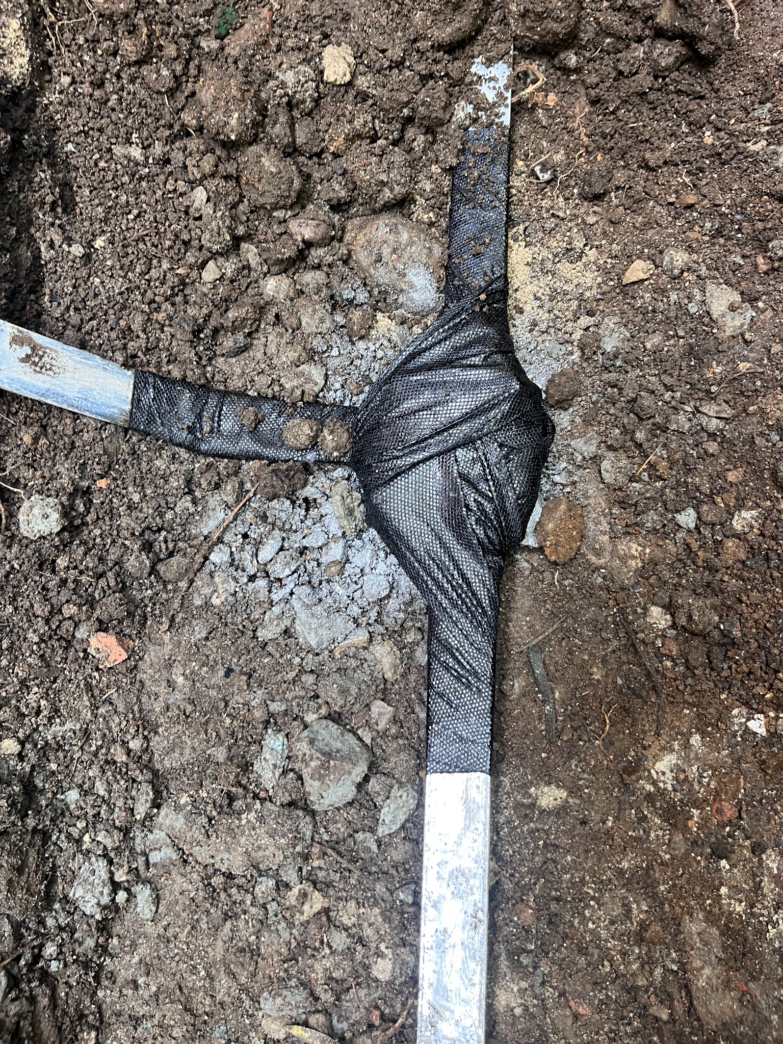 Uziom otokowy w ziemi, zabezpieczony czarną siatką, widoczne kamienie i struktura gruntu. Detal wykonawczy trasy zasilania budynku w trudnych warunkach geologicznych.