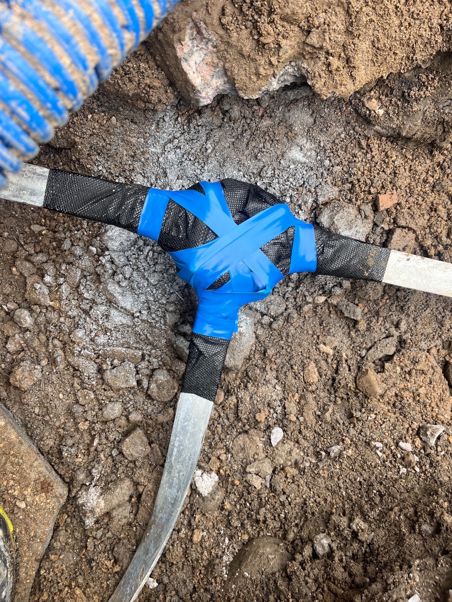 Połączenie trzech przewodów w ziemi, zabezpieczone czarną taśmą i niebieską izolacją, widoczne z góry, w otoczeniu ziemi i gruzu.