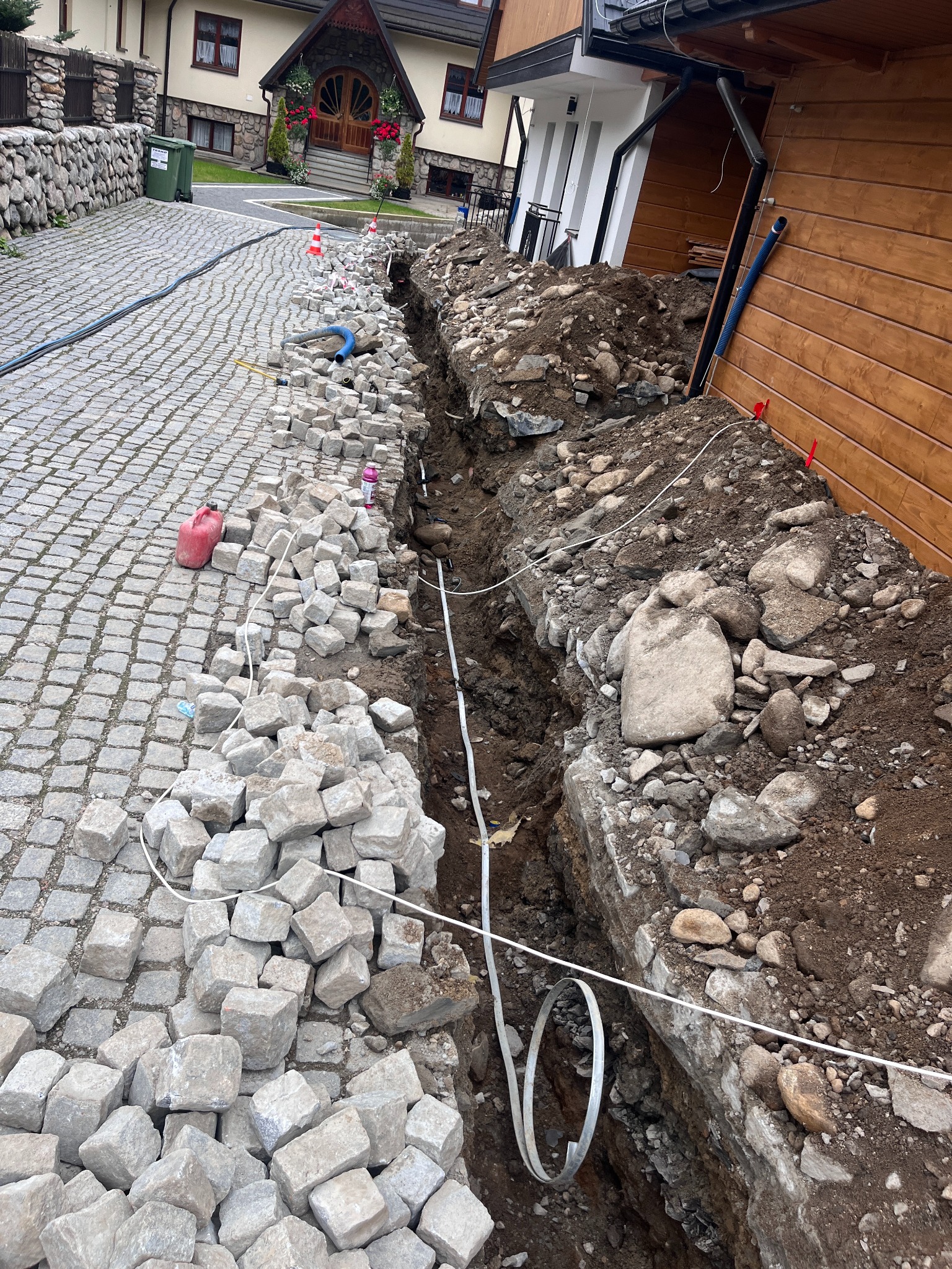 Wykop w ziemi z ułożonym przewodem. Obok kostka brukowa i narzędzia. Widok na budynek z drewnianą elewacją. Prace ziemne związane z instalacją.
