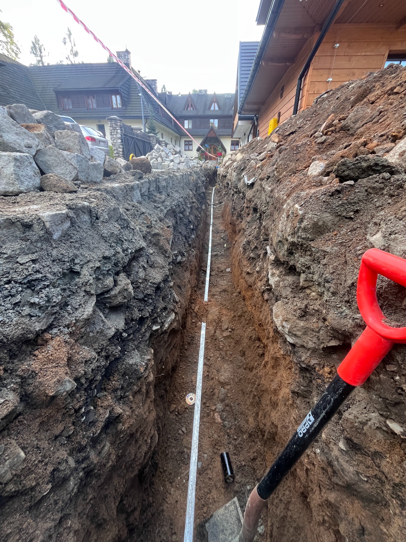 Wykop pod instalację elektryczną z ułożoną rurą osłonową, widoczna łopata z czerwoną rączką i budynek w tle. Prace ziemne przed zasypaniem.