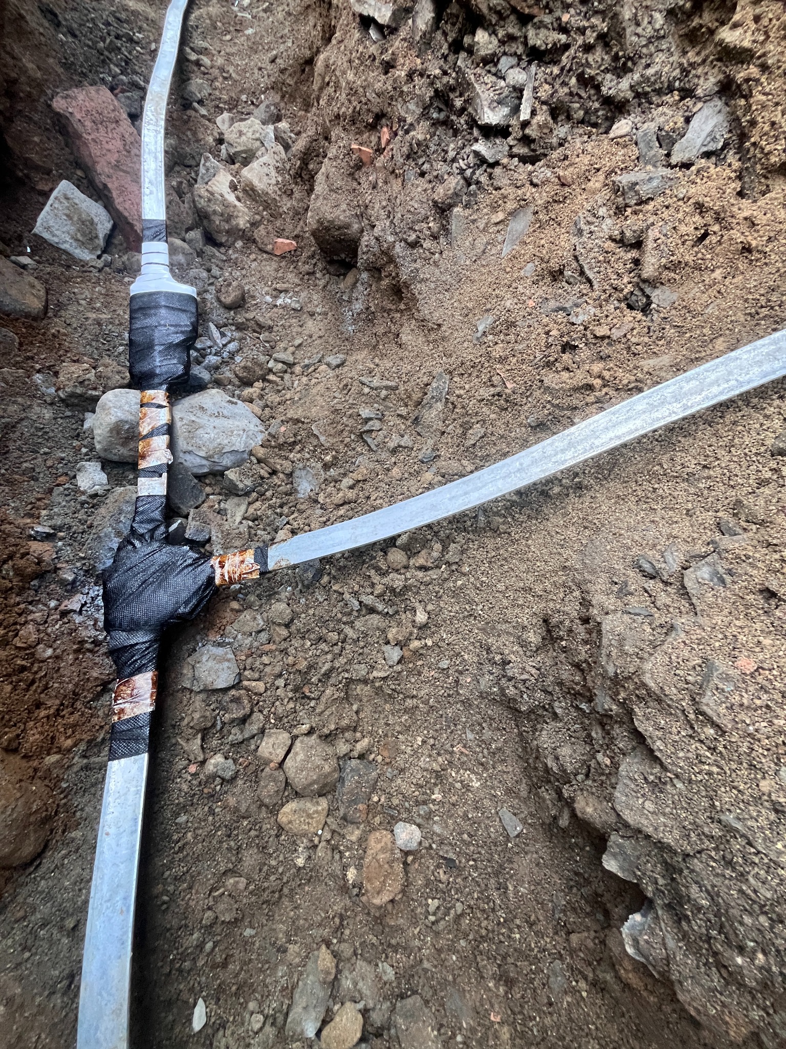 Metalowa rura w ziemi, zabezpieczona czarną taśmą izolacyjną, widoczna w wykopie z gruzem i ziemią. Element instalacji zakopany w ziemi.