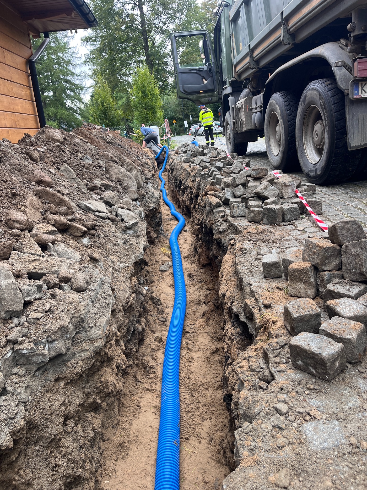 Niebieska rura karbowana w wykopie, prawdopodobnie pod kable, z pracownikami i ciężarówką w tle. Widoczne kostki brukowe i ziemia.