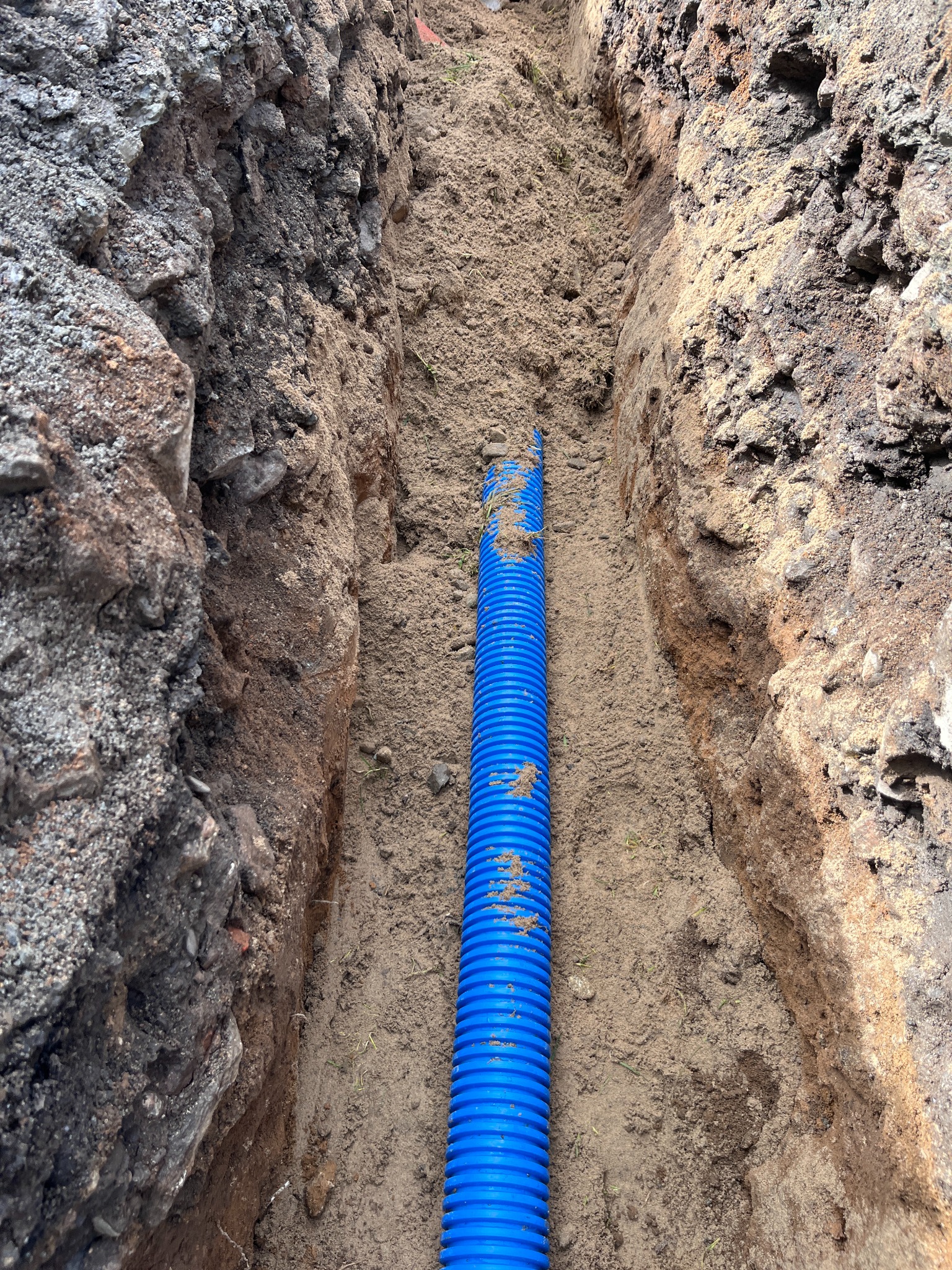 Niebieska rura karbowana w wykopie ziemnym, prawdopodobnie do kabli. Widoczne ślady ziemi na rurze. Wykop o nierównych ścianach z różną strukturą gleby.