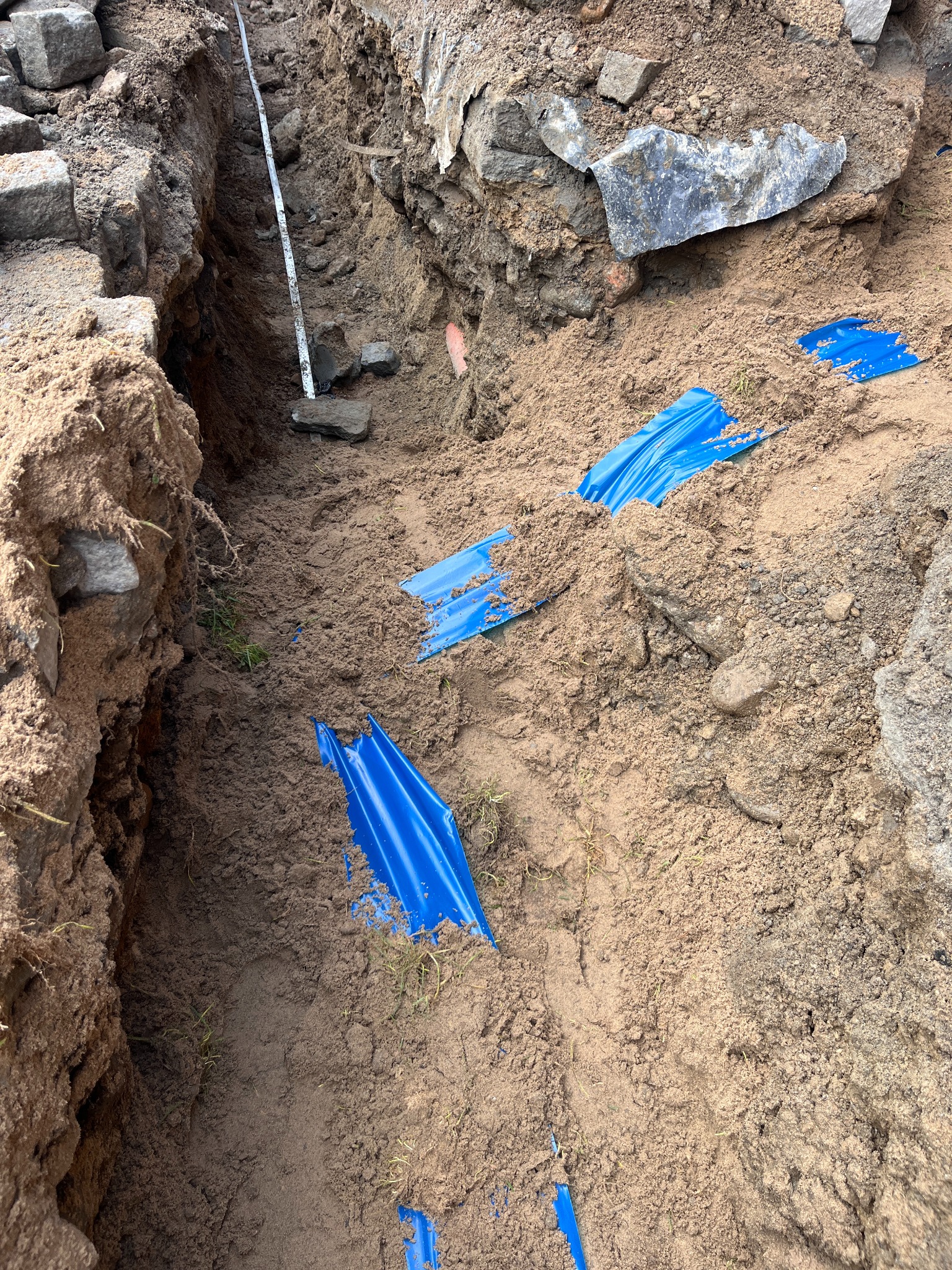 Wykop w ziemi z niebieską folią ochronną kabli, widoczny fragment miarki. Przygotowanie do ułożenia przewodów w ziemi.