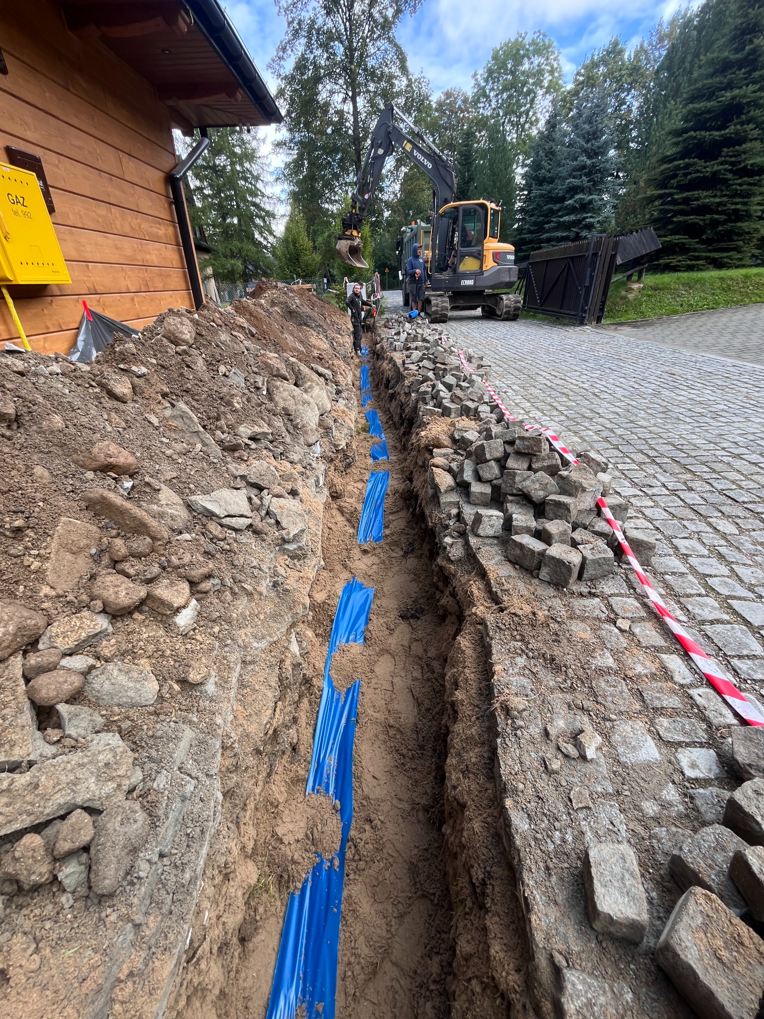Wykop z niebieskimi rurami w ziemi, przygotowany pod ułożenie kabli. Na górze koparka i kostka brukowa. Żółta skrzynka gazowa na drewnianym budynku w tle.