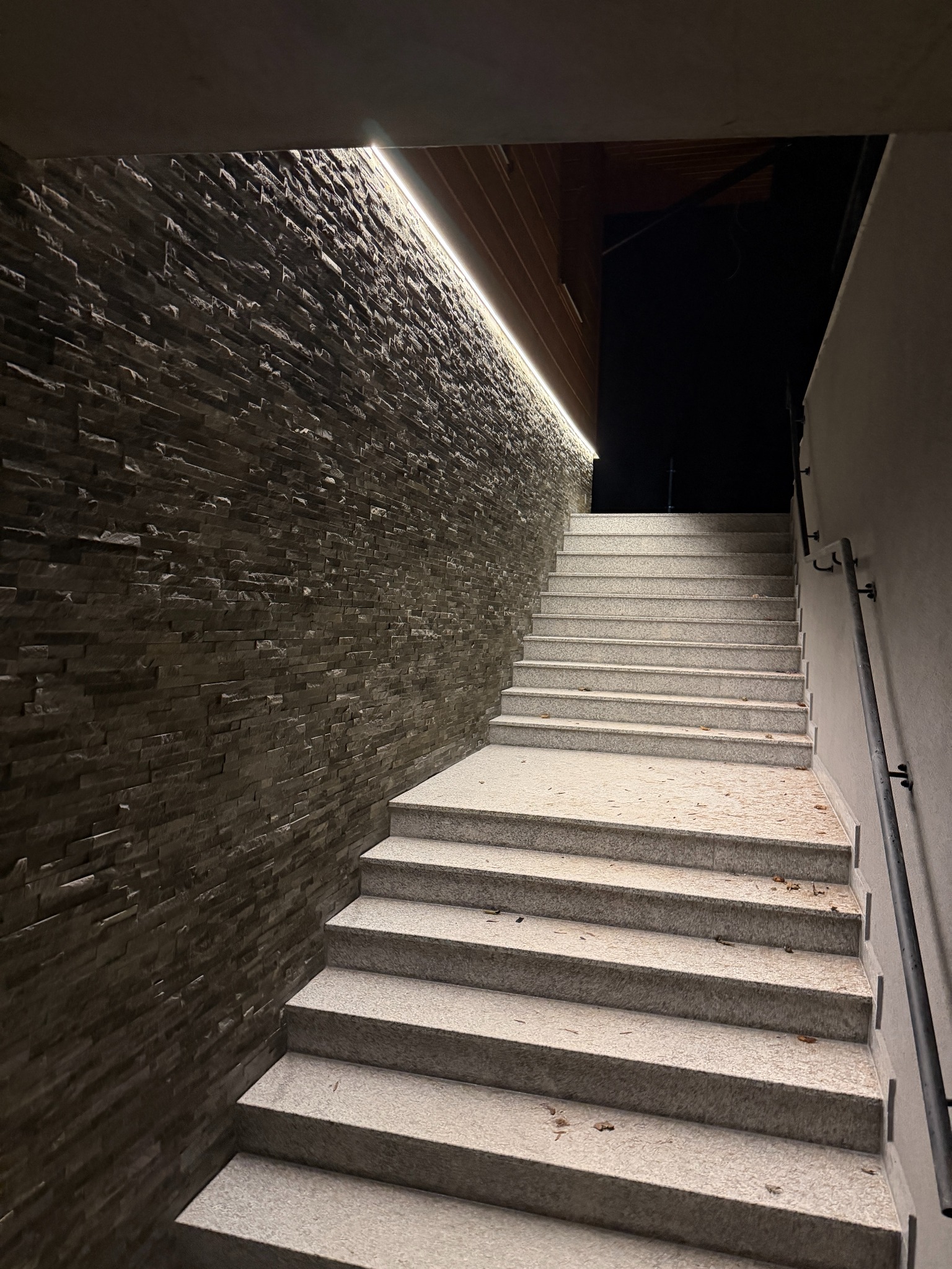 Oświetlone schody zewnętrzne z kamienną ścianą i taśmą LED pod balkonem, nocna elewacja. Rozbudowa oświetlenia zewnętrznego.