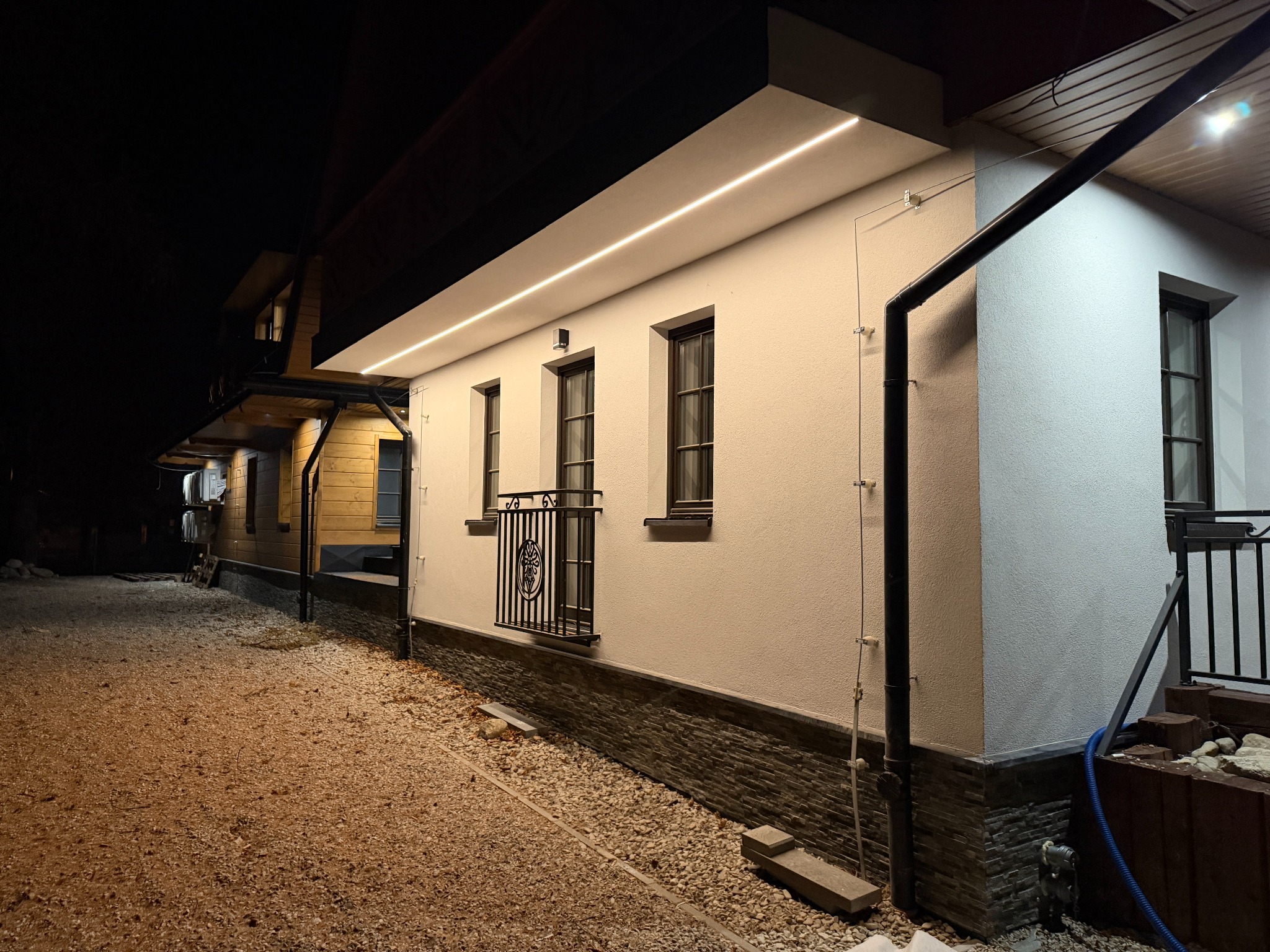 Oświetlenie elewacyjne LED: biały budynek z trzema oknami i balkonem, podświetlony liniowym światłem LED pod okapem, nocne ujęcie z żwirowym podłożem.
