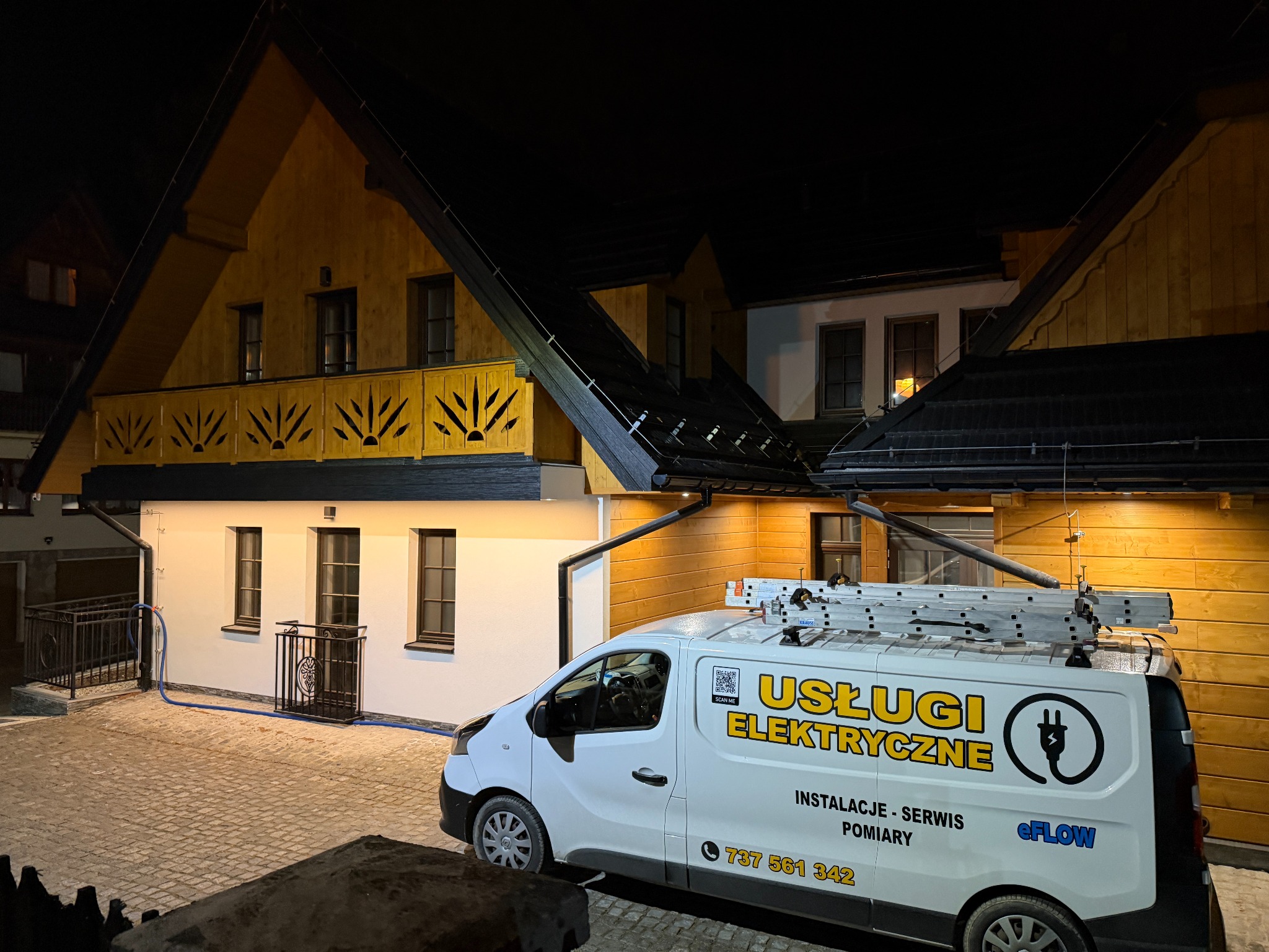 Biały van z napisem 'Usługi Elektryczne' zaparkowany przed domem w stylu góralskim nocą. Na dachu drabina, widoczny numer telefonu i logo firmy.