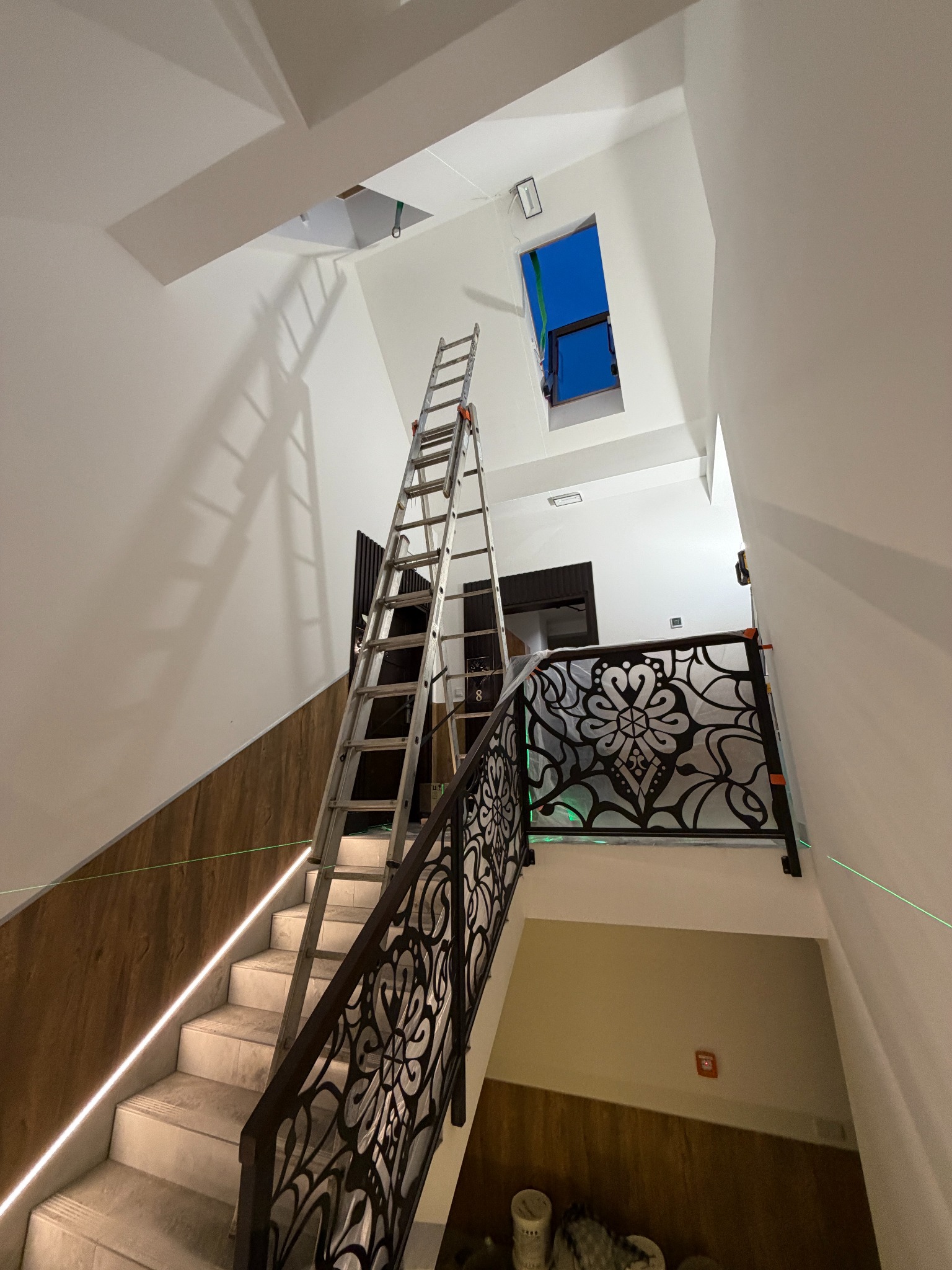 Klatka schodowa z drabiną, widoczne prace na wysokości. Laserowy poziom wskazuje na precyzyjne ustawienie. Montaż oświetlenia w trudnodostępnym miejscu.