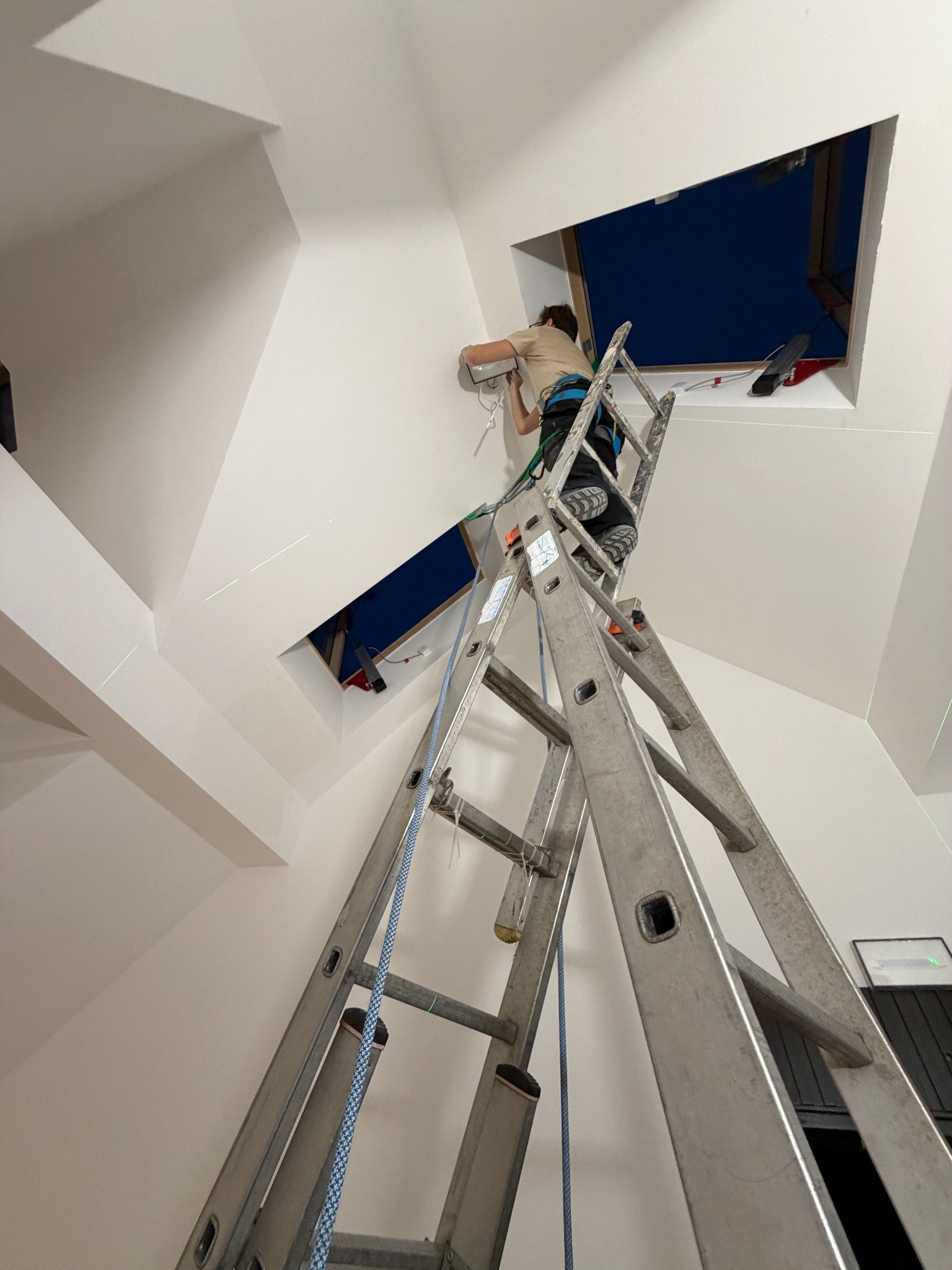 Elektryk na drabinie montuje instalację w otworze w skośnym suficie. Widoczne przewody i laserowy wskaźnik poziomu. Nietypowy kształt otworu okiennego.