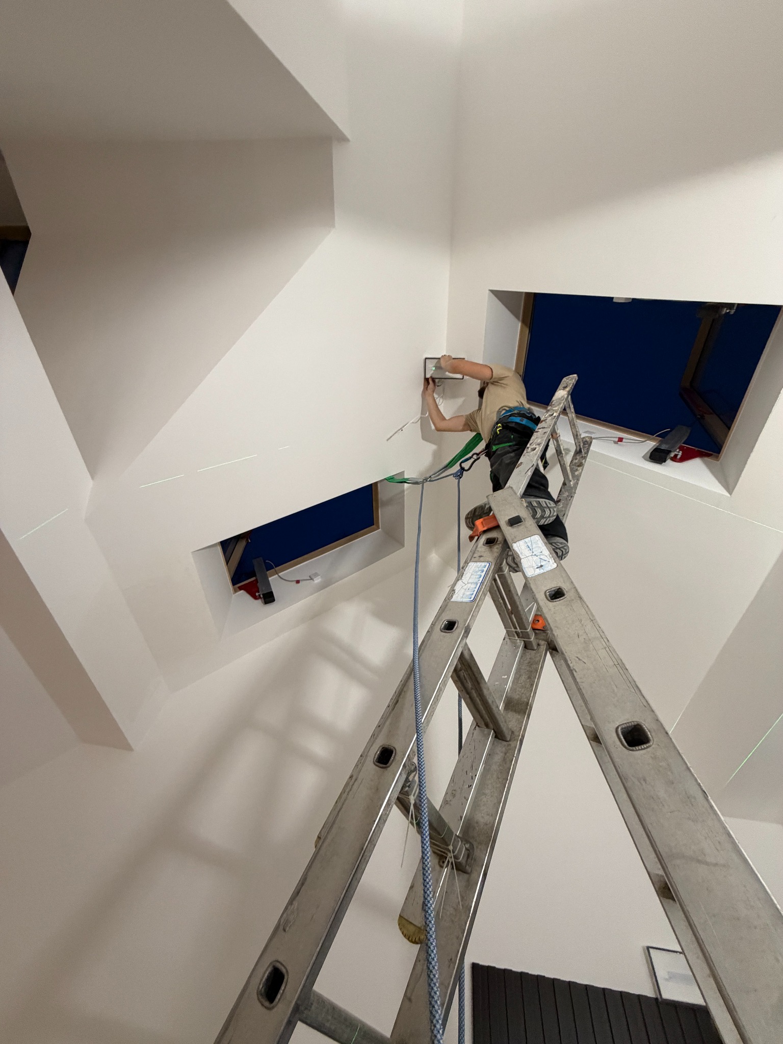Elektryk na drabinie montuje osprzęt w skosie pod oknem dachowym, zabezpieczony uprzężą. Widoczne zielone linie lasera wyznaczające poziom. Białe ściany i sufit.