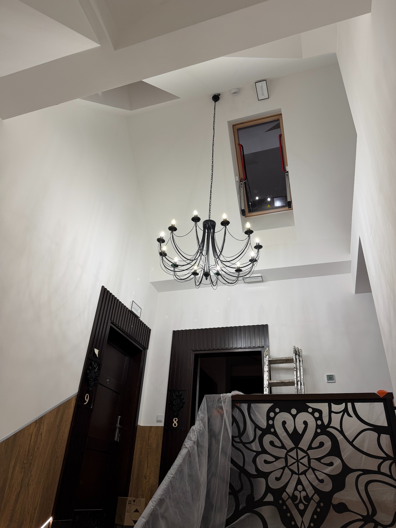 Nowoczesny żyrandol w jasnym holu z wysokim sufitem i oknem dachowym, widoczne drzwi z numerami mieszkań i fragment metalowej balustrady.