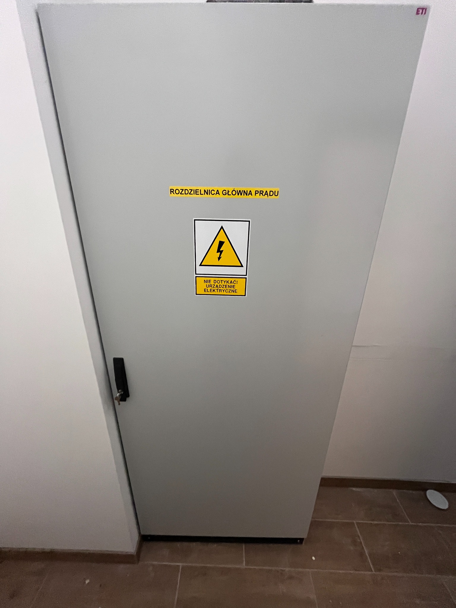 Szara rozdzielnica główna prądu z żółtą naklejką ostrzegawczą o napięciu, umieszczona w rogu pomieszczenia z brązową podłogą.