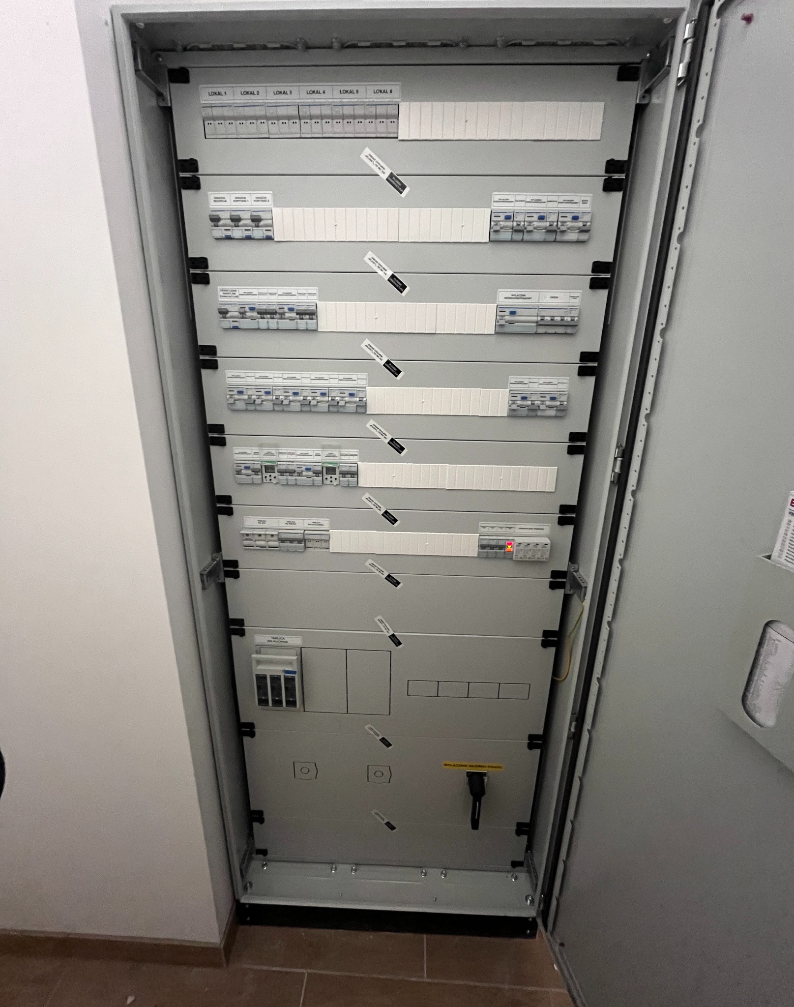 Otwarta szafa rozdzielcza z bezpiecznikami i oznaczeniami lokalizacji, widok z góry, szara obudowa, instalacja elektryczna wewnątrz budynku.