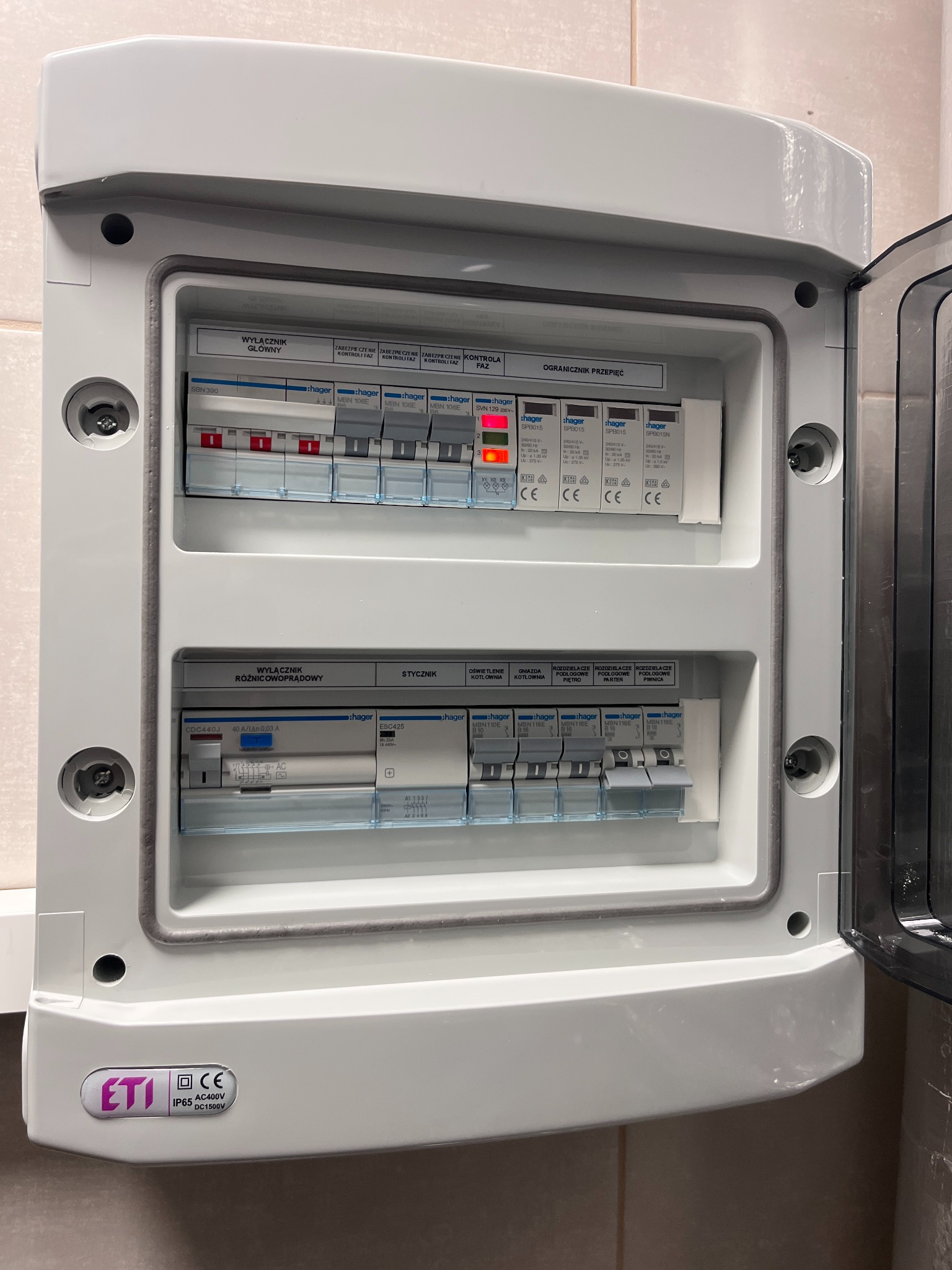 Otwarta, biała skrzynka rozdzielcza z widocznymi bezpiecznikami i wyłącznikami. Widoczne oznaczenia producentów Hager i ETI. Jeden z bezpieczników sygnalizuje awarię.
