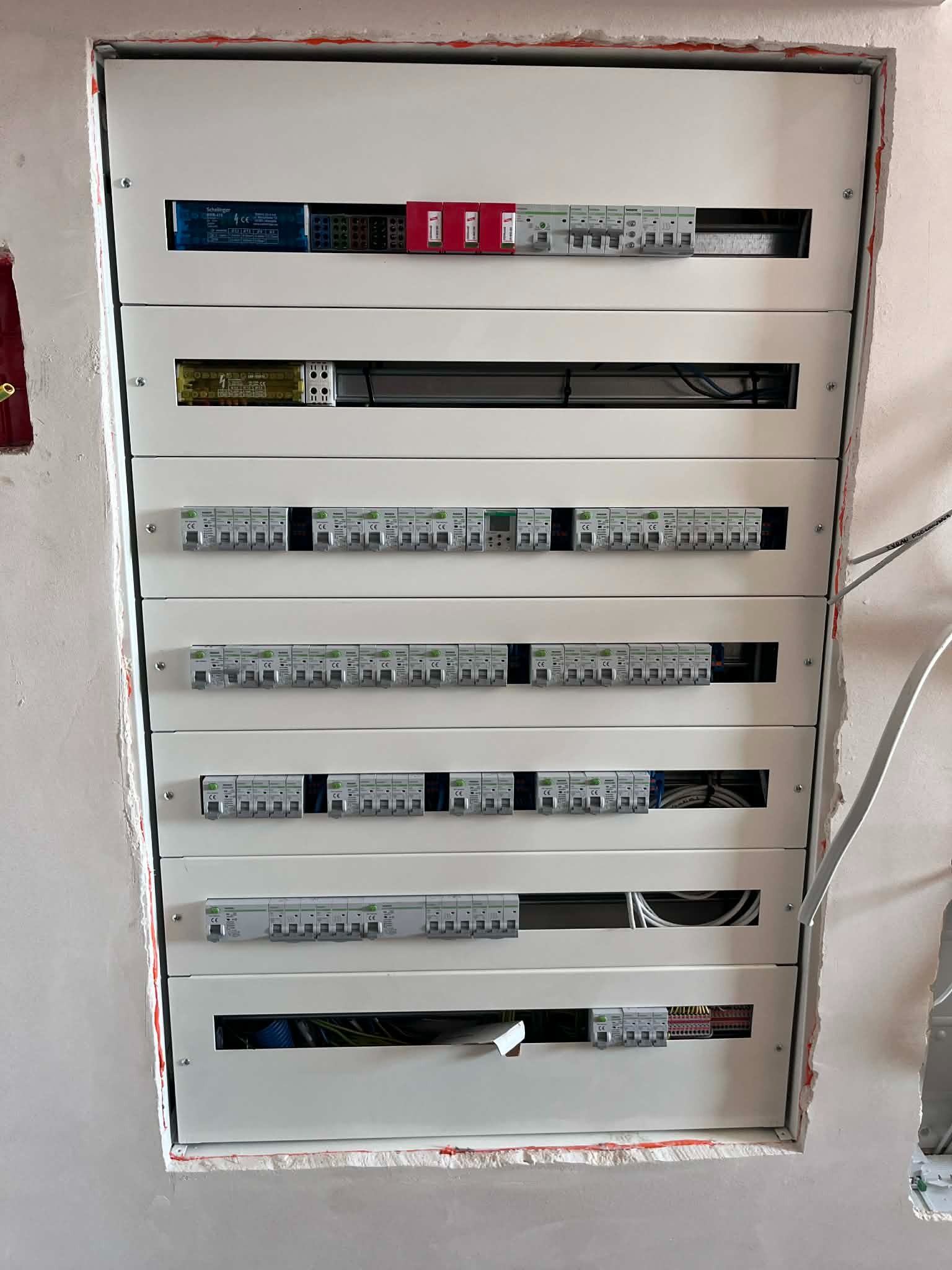 Rozdzielnica elektryczna wnękowa, z licznymi zabezpieczeniami i przewodami, zamontowana w surowej ścianie. Widoczne rezerwy na przyszłą rozbudowę.