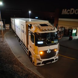 Trans-Bus Konrad Koza - Biały samochód ciężarowy Mercedes-Benz Actros z nadrukiem Trans-Bus, zaparkowany nocą na parkingu przed budynkiem McDonald's i Bricoman.