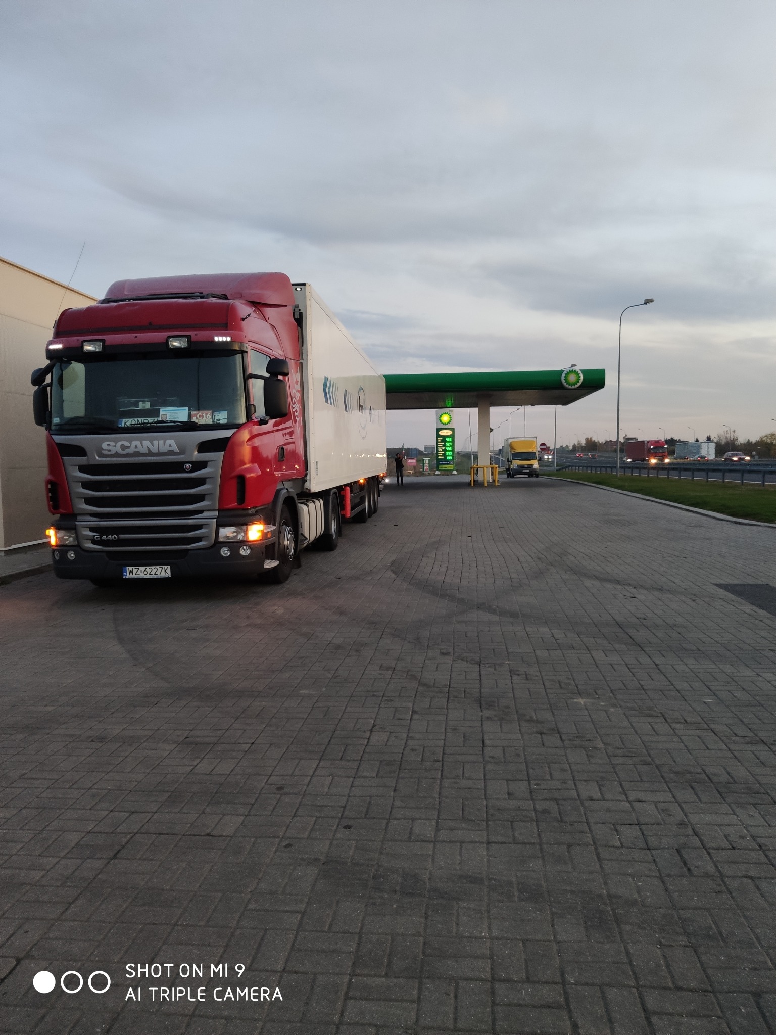 Czerwona ciężarówka Scania G440 z białą naczepą na stacji benzynowej BP, widok z przodu, tablica rejestracyjna WZ 6227K, szara kostka brukowa, pochmurne niebo.