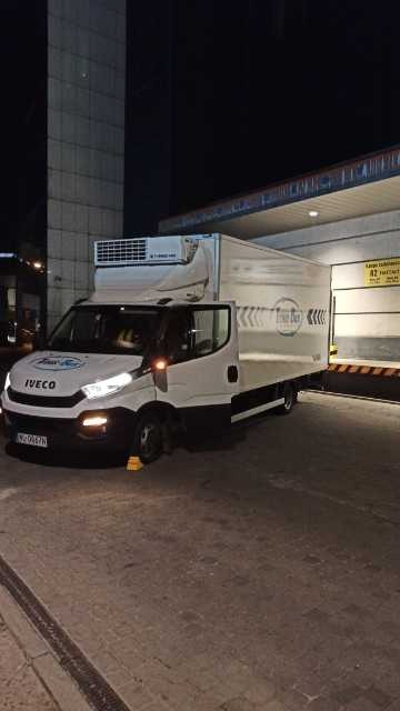 Biały bus chłodnia marki Iveco, zaparkowany nocą przy rampie załadunkowej, z włączonymi światłami. Żółte kliny pod kołami. Widoczne logo firmy transportowej.