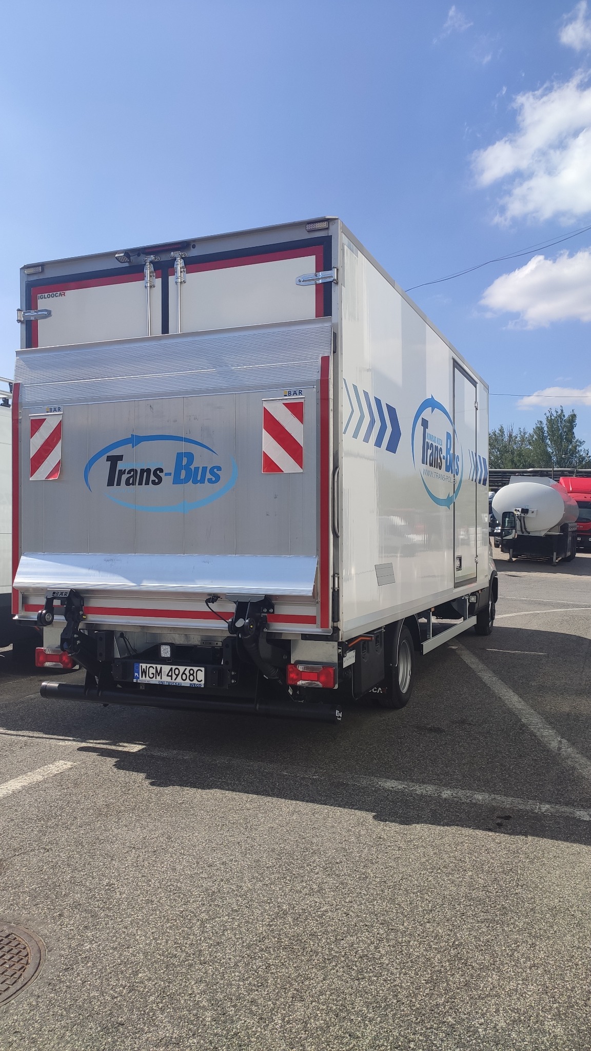 Tył białego busa transportowego z windą załadowczą na parkingu w słoneczny dzień. Widoczne logo firmy i tablica rejestracyjna.