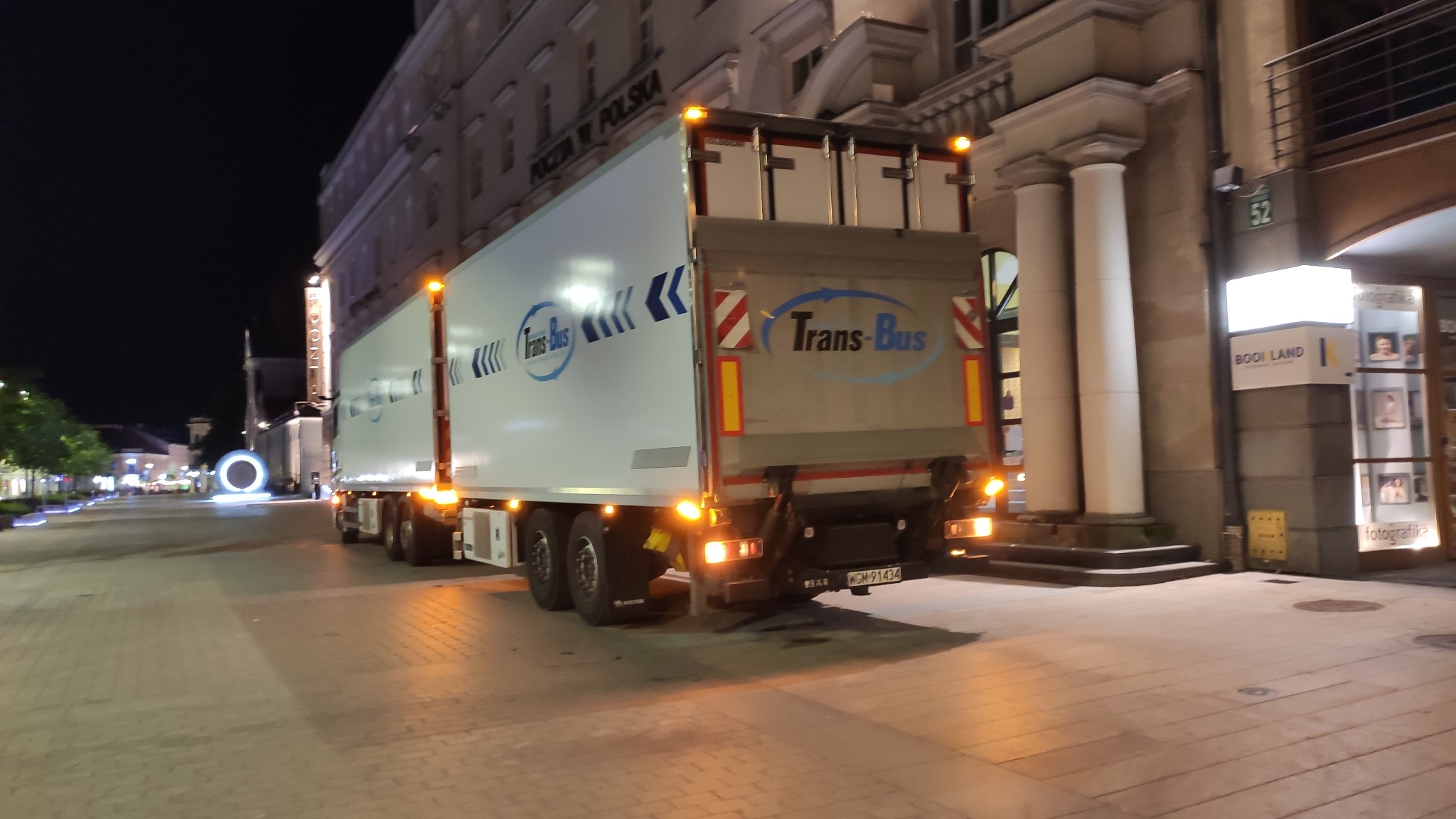 Ciężarówka z naczepą marki Trans-Bus zaparkowana nocą na ulicy miasta, z widocznym budynkiem Poczty Polskiej w tle i oświetlonymi dekoracjami miejskimi.