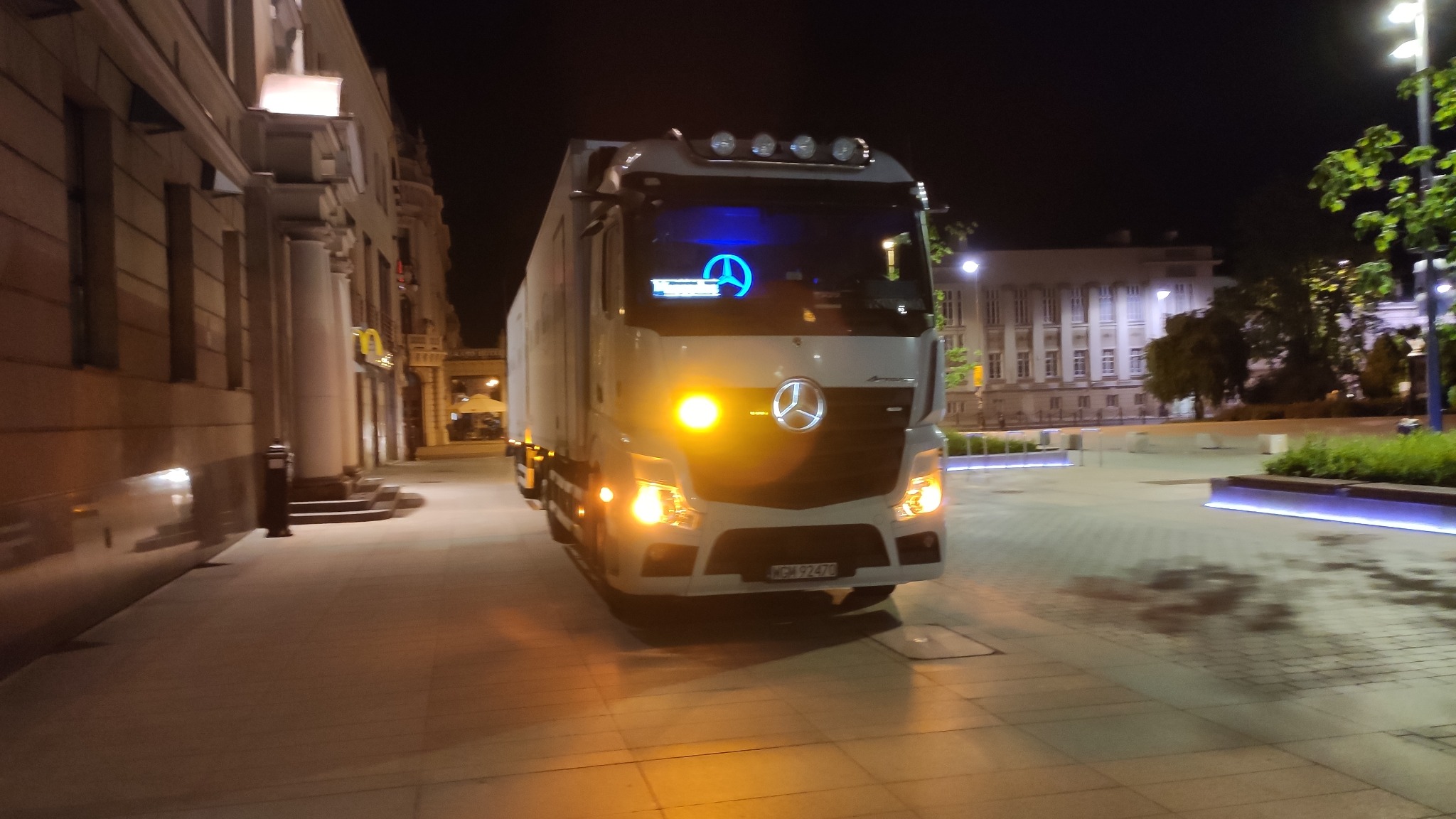 Biały bus Mercedes z niebieskim logo na szybie, nocą na tle architektury Warszawy. Światła pojazdu odbijają się od chodnika.