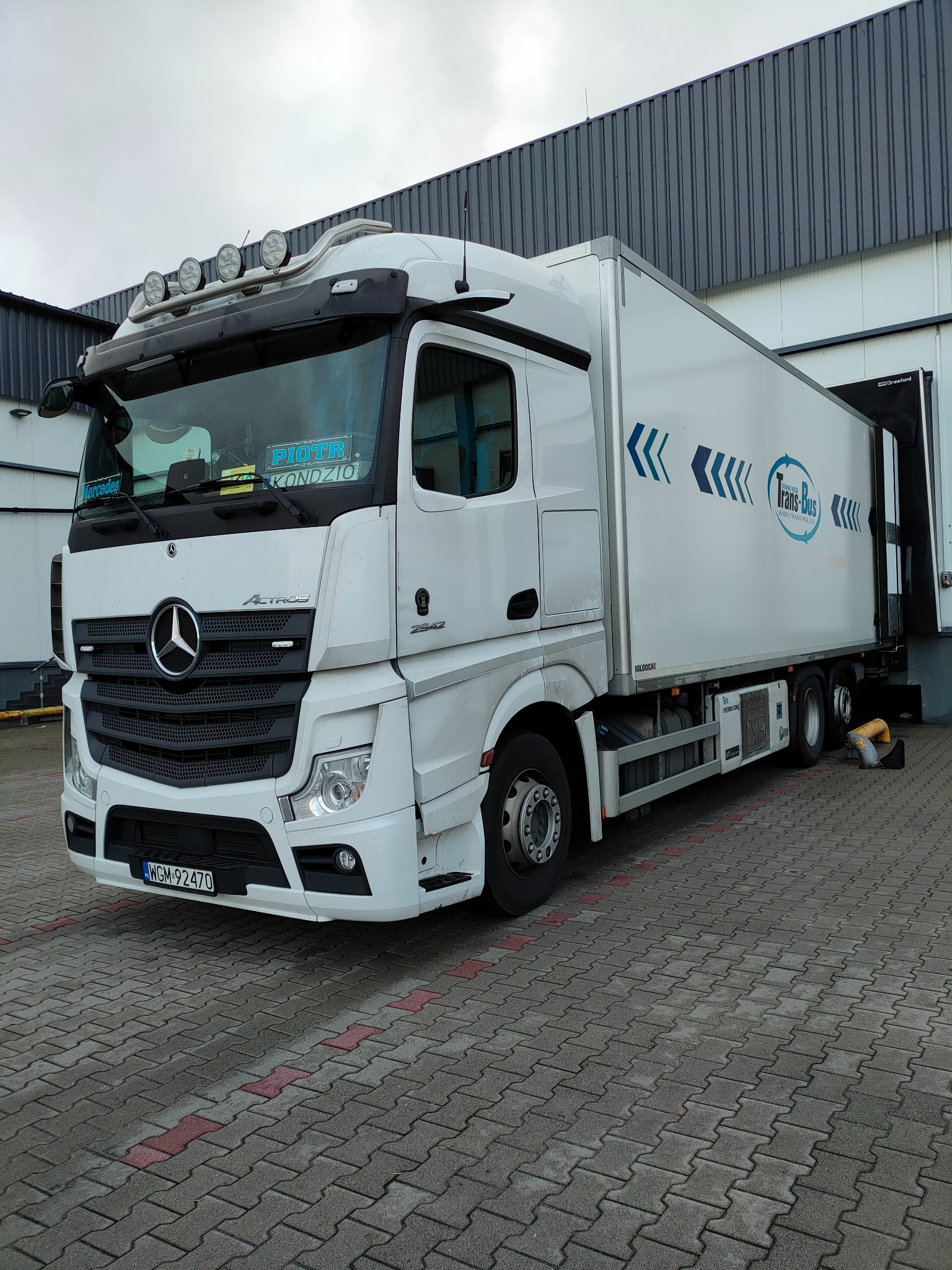 Biały Mercedes Actros z naczepą Trans-Bus, zaparkowany na kostce brukowej przed budynkiem magazynowym z rampą. Widoczne dodatkowe lampy na dachu kabiny.
