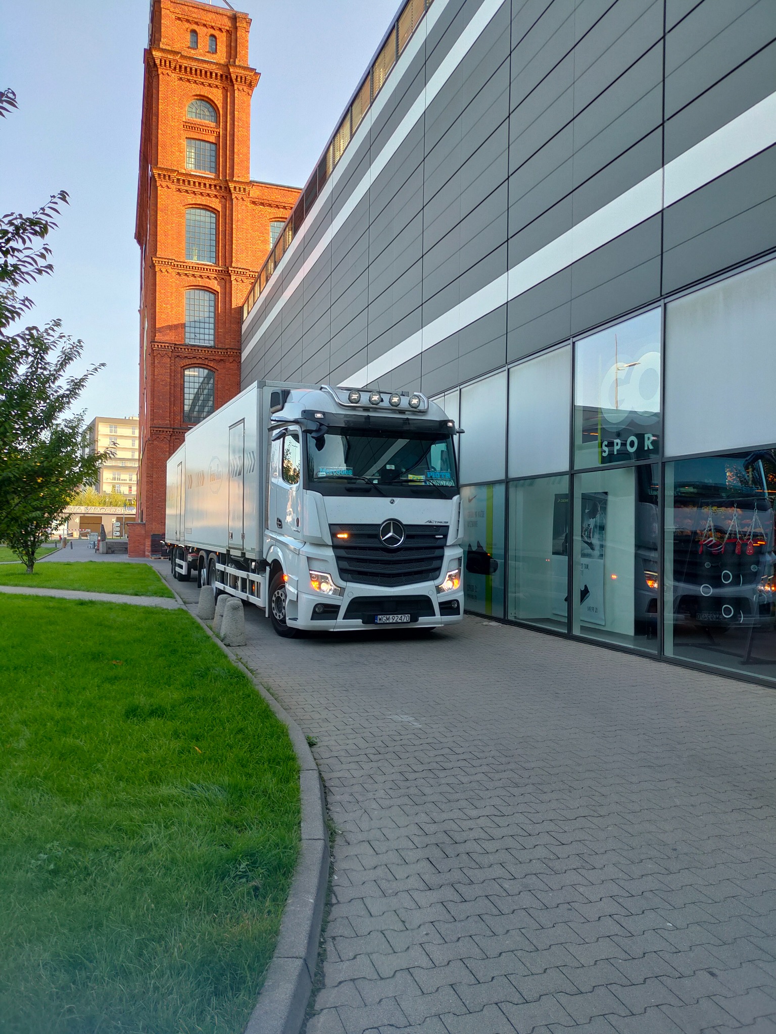Biały samochód ciężarowy Mercedes-Benz Actros z naczepą na tle ceglanej wieży i nowoczesnego budynku ze szklanymi witrynami.