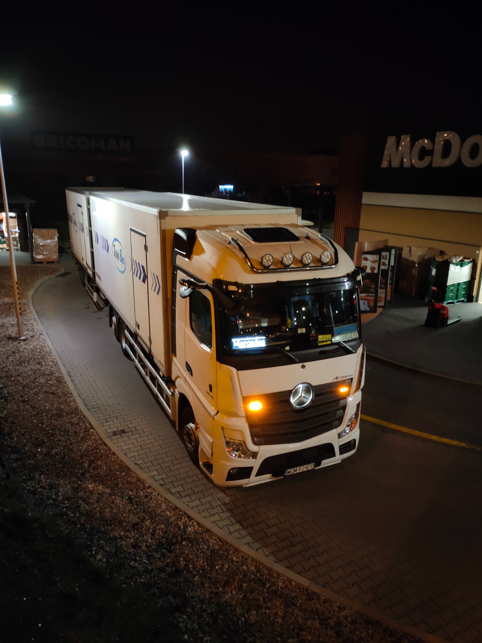 Biały samochód ciężarowy Mercedes-Benz Actros z nadrukiem Trans-Bus, zaparkowany nocą na parkingu przed budynkiem McDonald's i Bricoman.