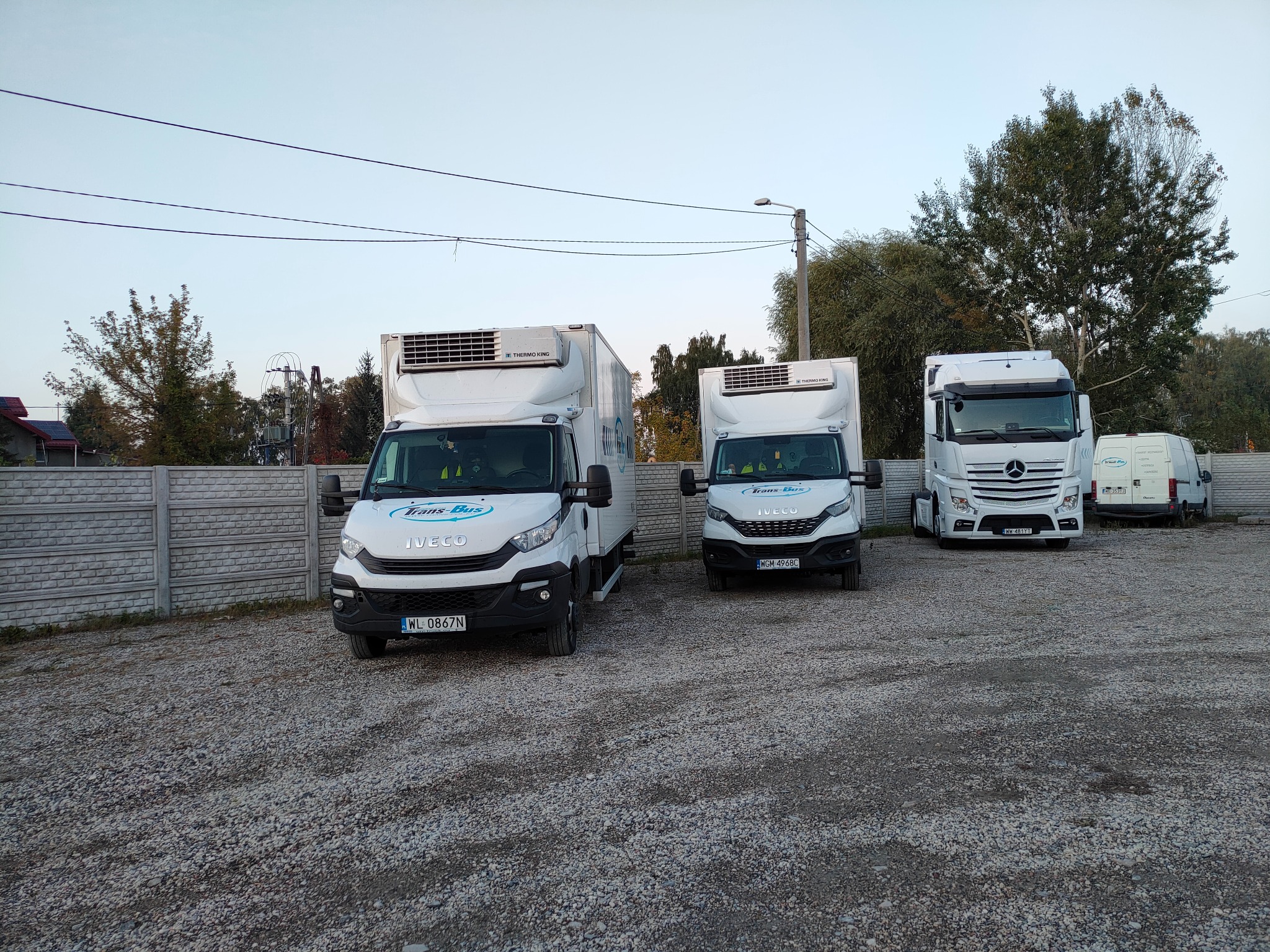 Flota białych pojazdów dostawczych Iveco i Mercedes z nadrukiem 'Trans-Bus' na żwirowym placu, w tle betonowe ogrodzenie i drzewa pod błękitnym niebem.